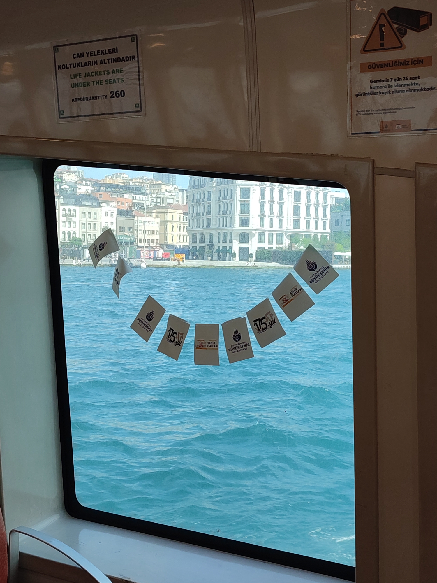 This is an interior shot taken from a ferry crossing the Bosphorus Strait, likely near the Beyoğlu district of Istanbul, Turkey. Through a window, the bright blue, choppy waters of the strait are visible, with a city skyline on the far bank featuring numerous buildings, some with balconies and white facades.
Inside the ferry, adhered to the window, are several small white flags or pennants. Some of these appear to be promotional or commemorative, bearing logos and text. Specifically, one flag shows the Istanbul Metropolitan Municipality logo and the text "İSTANBUL BÜYÜKŞEHİR BELEDİYESİ" (Istanbul Metropolitan Municipality). Another flag displays "75. YIL" (75th Year), suggesting a potential anniversary commemoration. There are also flags with text that is partially obscured or illegible.
On the upper interior wall of the ferry, to the left, is a sign in both Turkish and English stating "CAN YELEKLERİ KOLTUKLARIN ALTINDADIR LIFE JACKETS ARE UNDER THE SEATS ADED/QUANTITY 260". To the right, another sign features an orange triangular warning symbol with a camera inside, and text in Turkish indicating that the ferry is monitored by cameras 24/7 for security.
The overall scene depicts a common mode of public transport in Istanbul, with practical safety information displayed alongside decorative elements. No people are clearly visible in the image.