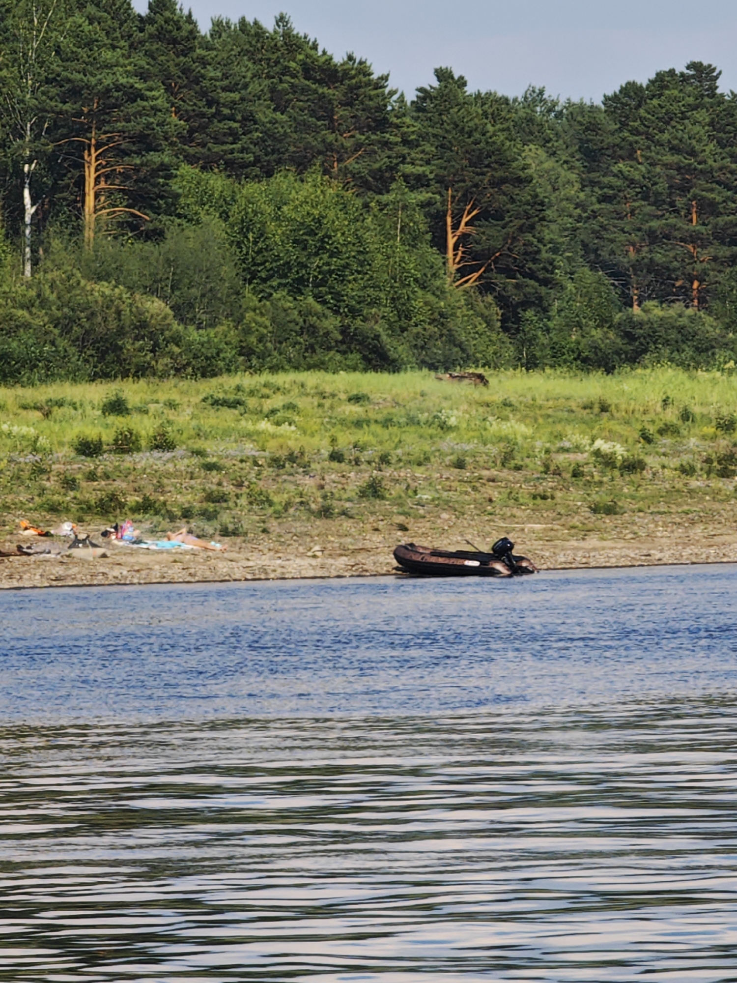 This image depicts a serene natural scene by the river in Prutovaya, Russia. In the foreground, the wide expanse of a blue river with visible ripples stretches across the frame. The middle ground reveals a sandy and pebbly riverbank dotted with sparse vegetation. Several people are seen sunbathing on towels on the riverbank. An inflatable boat with an outboard motor is moored near the shore. The background is dominated by a dense forest with a mix of deciduous and coniferous trees, and beyond that, a clear blue sky. The overall impression is one of a tranquil summer day in a rural Russian setting.