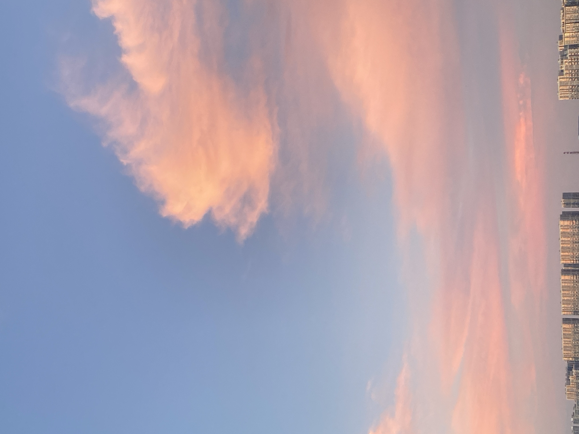 A cityscape is visible in the distance under a vast sky filled with soft pink and orange clouds at sunset. The sky transitions from a light blue at the top to warmer hues near the horizon, with the pink clouds dominating the mid-to-upper portion of the frame. Below the sky, a strip of buildings stretches horizontally across the bottom of the image. These buildings appear to be apartment complexes or similar multi-story structures, with many windows visible. The overall impression is one of a peaceful urban sunset.