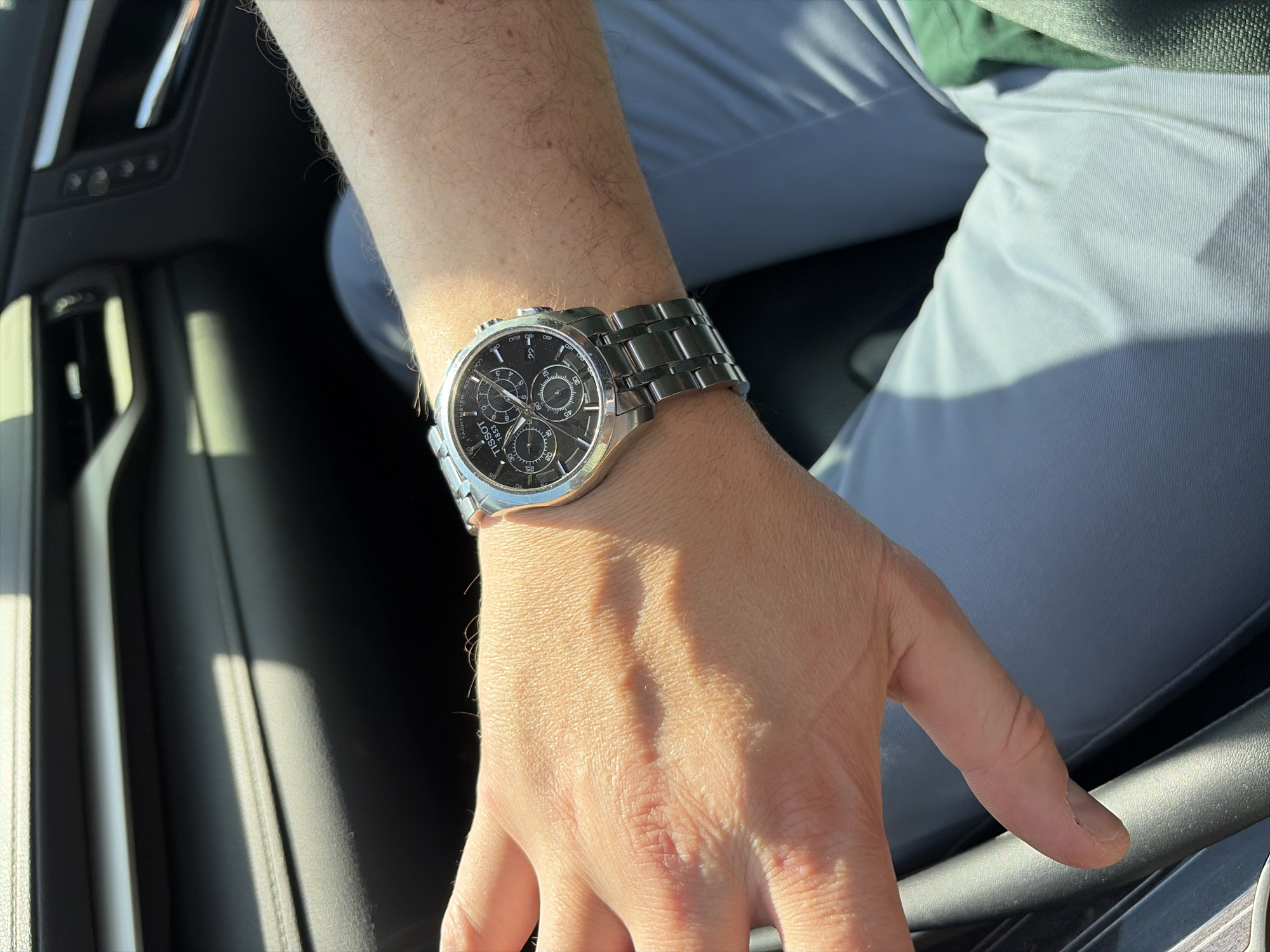 The image shows a close-up of a person's left wrist and forearm, wearing a silver Tissot chronograph watch with a black dial. The person's hand is visible, resting on a dark, textured surface that appears to be the interior of a car, possibly a dashboard or seat. Sunlight is casting strong shadows across the hand and arm, indicating a bright, sunny day. The background is out of focus, showing the dark interior of the car, including what looks like a car door handle and air vent. The person is wearing light blue pants. The context does not provide any information about the location being Moscow, Russia.