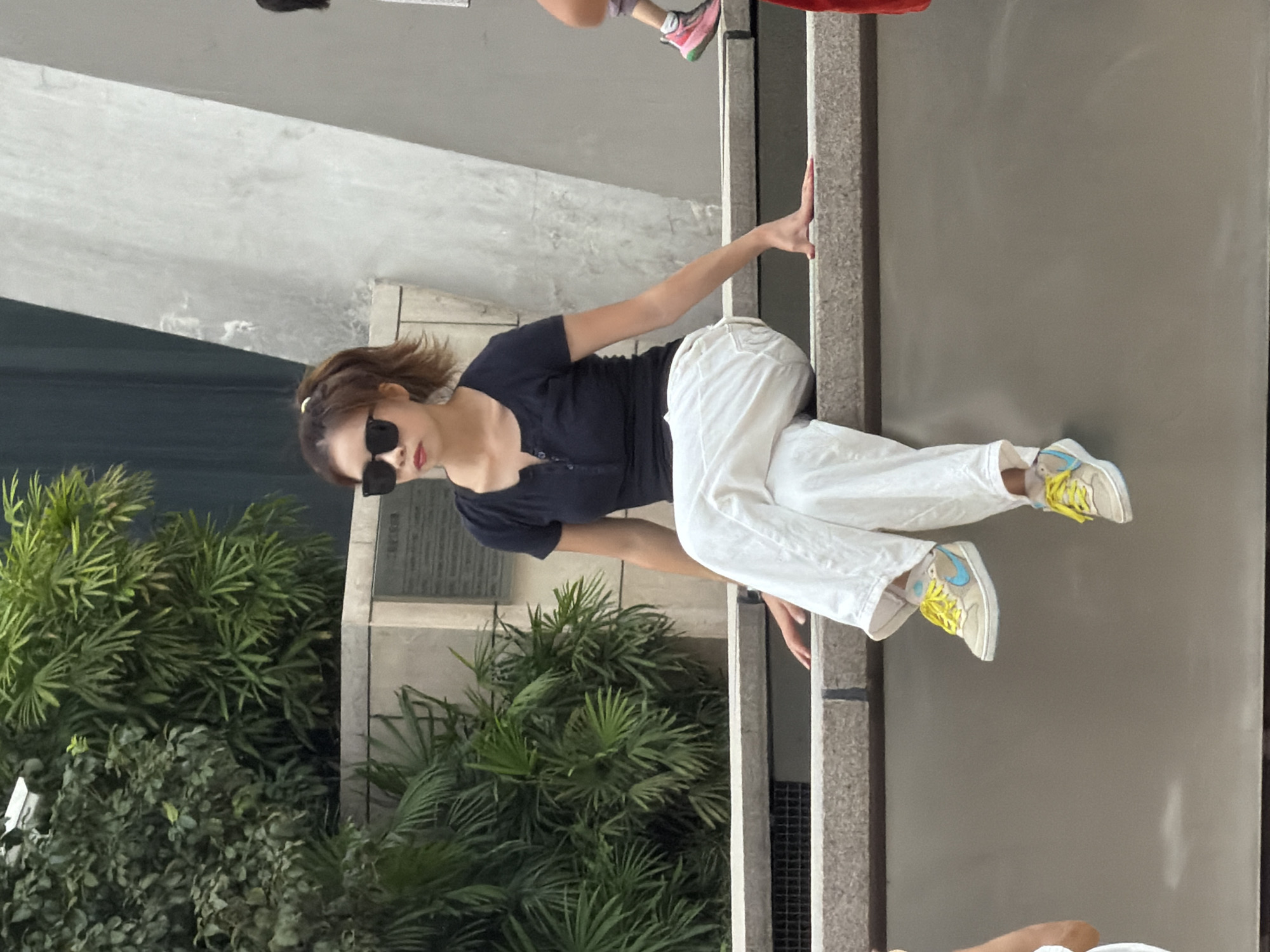 A young Asian woman with brown hair tied in a ponytail is sitting on a concrete ledge outdoors, wearing a black short-sleeved shirt, white pants, and tan sneakers with blue accents and yellow laces. She has on black sunglasses and is looking to her left. The environment includes lush green foliage and a grey concrete structure behind her. Another person's legs wearing pink athletic shoes and purple pants are visible to her right. This scene is located in Yuzhong District, China.