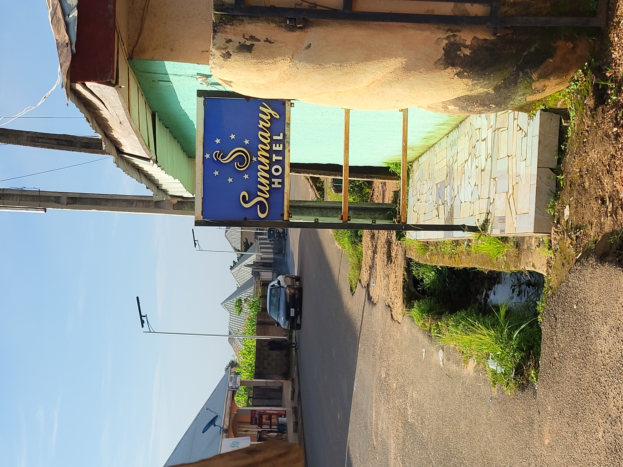 The image depicts a street scene in Barakin Akawo, Nigeria. In the foreground, a weathered, light brown, rounded structure, possibly a decorative element or part of a building, stands next to a black metal fence. A blue sign with a golden "S" logo and the words "Summary HOTEL" in golden script is prominently displayed on a metal pole. The sign is bordered by small golden stars. A paved walkway, made of irregularly shaped light-colored tiles, leads away from the sign. To the left of the walkway, a narrow, overgrown ditch with murky water runs alongside the road.
The road itself is asphalt and appears to be a two-lane street. A dark-colored sedan is driving away from the camera on the right side of the road. Further down the street, several residential buildings with distinctive pitched roofs are visible. A lone individual in dark clothing stands near a low brick wall on the left side of the road. On the far left, a building with a silver roof has a satellite dish and signage, suggesting a commercial establishment. The sky is clear and blue, indicating daylight. Vegetation, including tall grasses and some corn stalks, is present along the edges of the road and between buildings. The overall environment suggests a somewhat rural or semi-urban setting.