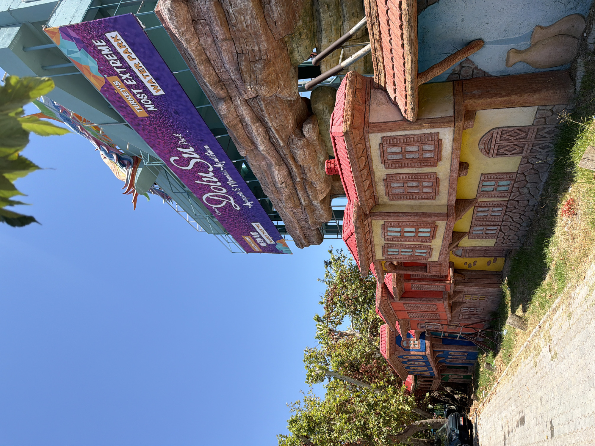 This is a full, eye-level, outdoor shot on a sunny day in Kemer, Turkey. The photograph features a vibrant and colorful row of buildings, styled to resemble traditional Turkish architecture, painted in shades of red, orange, yellow, and blue. These buildings are low-rise and appear to be decorative, possibly part of an amusement park or tourist attraction. Behind these buildings, a large, modern structure is visible, adorned with a prominent purple banner that reads "DoluSu Park" and "Turkey's Most Extreme Water Park." A portion of a colorful dragon sculpture, likely a ride or attraction, is also visible on this structure. The foreground shows a paved walkway with scattered grass and some debris. A black car is parked on the left side of the walkway. Trees with green foliage are present on the left and in the background, providing shade. The sky is a clear, bright blue.