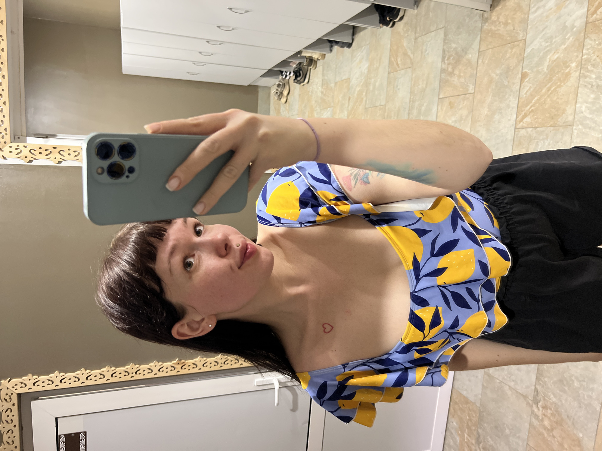 A young woman is featured in a mirror selfie, holding a light green smartphone with her right hand, capturing her reflection. She has dark hair with bangs and is looking towards the camera with a neutral expression, showing a slight pout. She is dressed in a light blue top featuring a cheerful pattern of yellow lemons and dark blue leaves, complemented by black shorts.
Distinctive personal details include a small red heart tattoo situated on her left collarbone and a faint, partially visible blue and pink tattoo on her right upper arm. She also has a small stud piercing above her upper lip and delicate stud earrings. A slender, light purple bracelet adorns her right wrist.
The indoor setting, identified as Chaikovsky, Russia, shows white drawers or cabinets with silver handles in the background, some of which display shoes on shelves. To the left, an intricately carved, light-colored decorative mirror frame adds to the room's decor. The floor is covered with light-toned, rectangular tiled flooring. The lighting suggests an interior, possibly artificially lit. No discernible text is visible in the image.