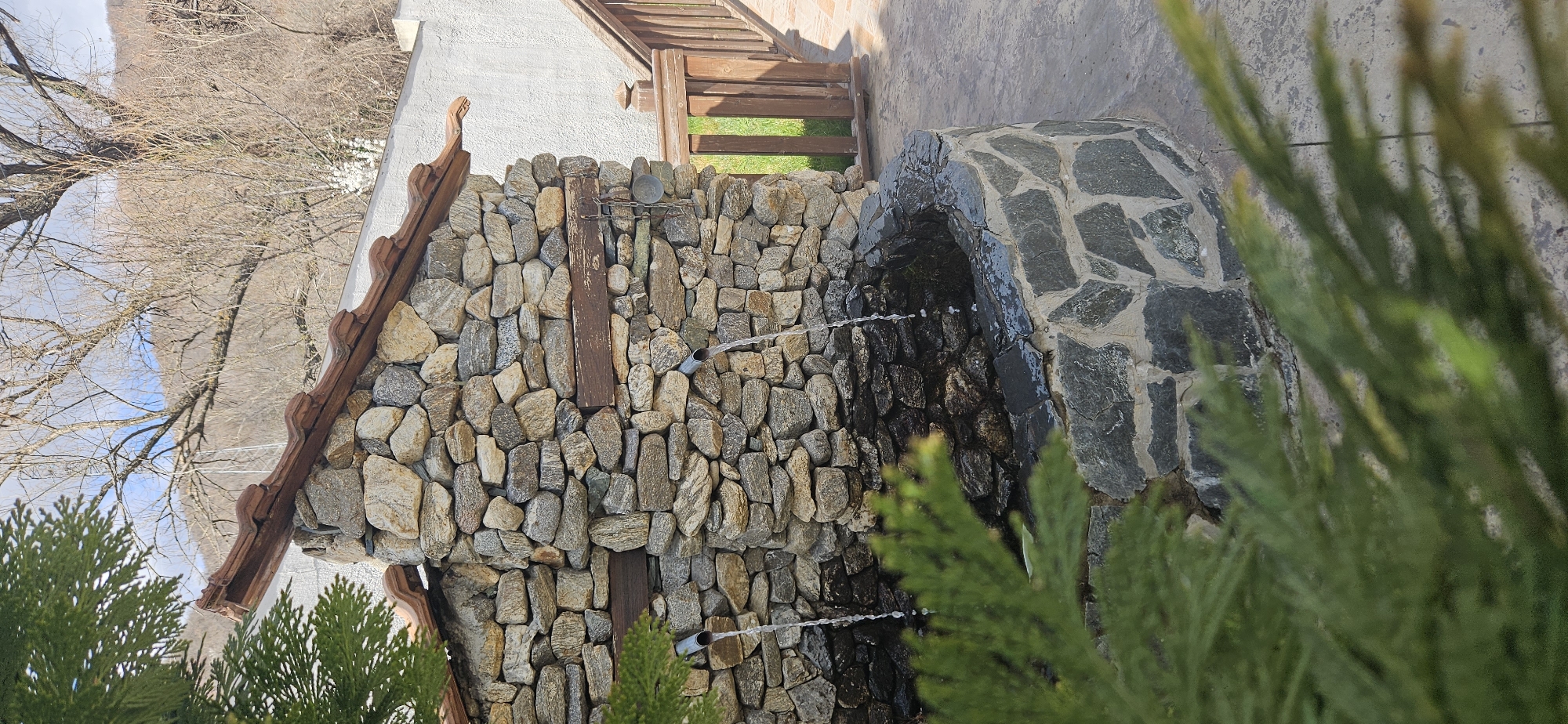 The image displays a stone-built fountain structure located in Rila, Bulgaria. The primary structure is a vertical wall constructed from irregularly shaped, light-colored natural stones, horizontally intersected by two dark wooden beams. Two metallic spouts extend from the stone wall below the beams, actively emitting streams of water into a semi-circular basin. This basin is fashioned from larger, dark-toned, irregularly shaped stones. A small, light-colored metal cup is suspended by a chain from the right-hand wooden beam. The top of the stone wall features a sloped roof made of reddish-brown clay tiles.
To the right of the fountain, a plain, light-colored stucco wall stands adjacent to a brown wooden railing, which borders a paved pathway leading towards a patch of green grass. The ground immediately surrounding the fountain is paved with light-grey flagstones or concrete. The background reveals bare deciduous trees and a distant, sparsely vegetated hillside under a partly cloudy sky. The lower-left foreground is partially obscured by green coniferous foliage. No individuals are present in the scene.