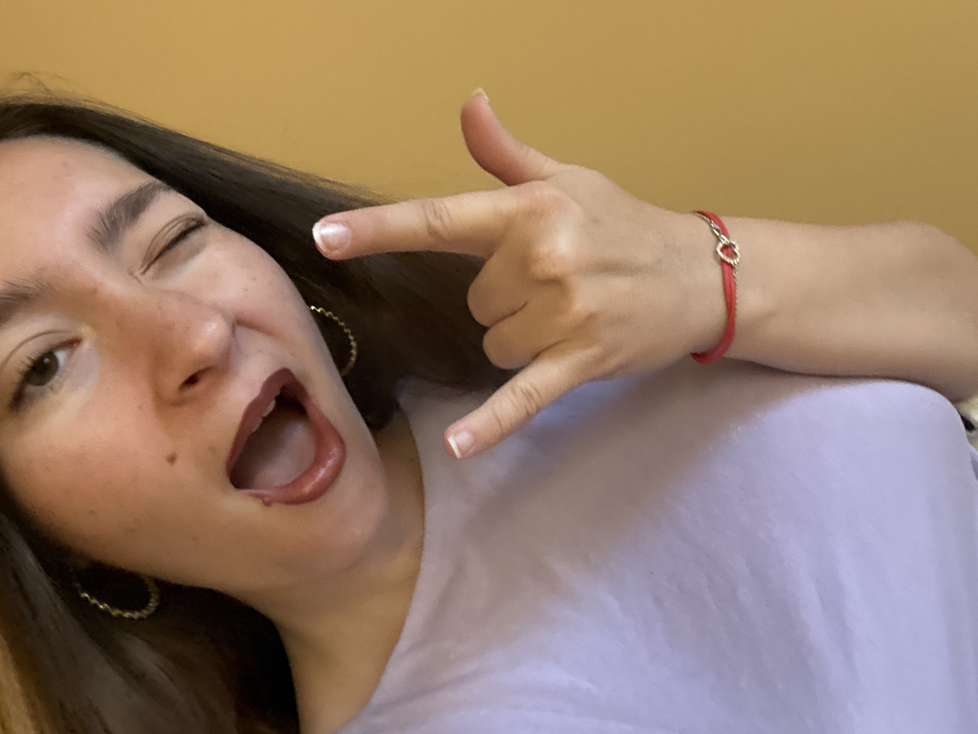 A young woman with dark hair is winking and sticking her tongue out slightly while holding up a "rock on" hand gesture. She is wearing a light purple top and several earrings. A red bracelet with a heart charm adorns her wrist. The background is a solid yellow wall. The image was taken indoors, possibly in a residential setting in Sofia, Bulgaria.