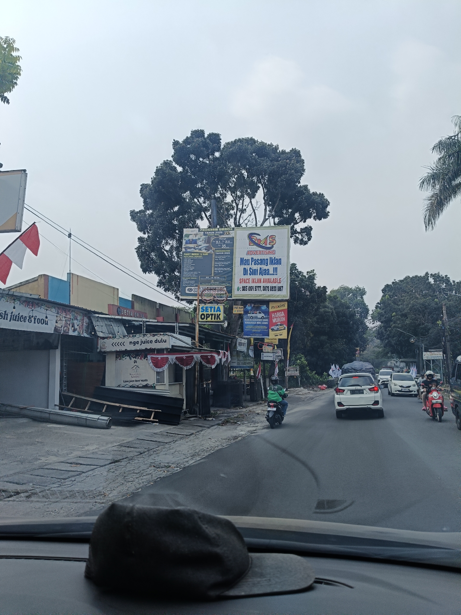 The image shows a street scene in Karaton, Indonesia. The foreground is dominated by the interior of a vehicle, with a dark fabric object resting on the dashboard. The view looks out onto a two-lane road with moderate traffic. Several cars and motorcycles are visible. On the left side of the road, there are buildings, including a shop with signage that reads "Nge juice dulu" and another with "Setia Optik." There are also several billboards, one advertising "SMS Advertising" and another for a "Program Umroh." The sky is overcast. The environment appears to be a commercial area with businesses lining the road.