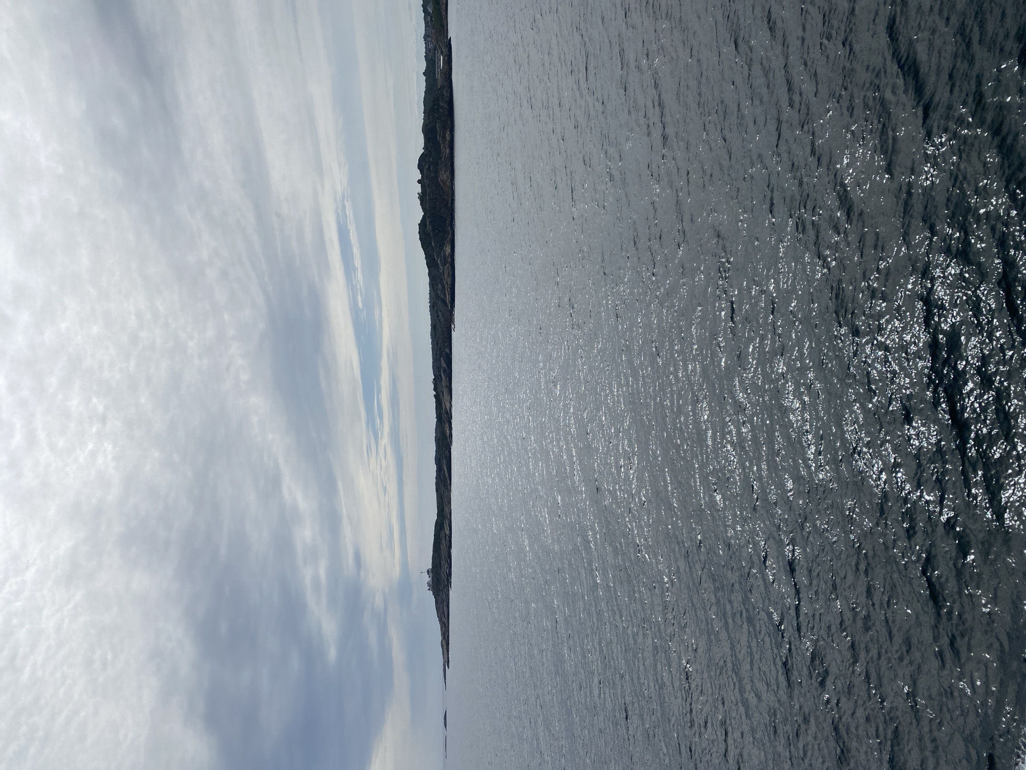 The image shows a vast expanse of dark, choppy water in the foreground, with sunlight reflecting off the surface in shimmering patterns. In the distance, a rocky, sparsely vegetated coastline stretches across the horizon. On the furthest point of the landmass, a white lighthouse with a red roof is visible. The sky above is overcast with a mottled pattern of grey and white clouds. The scene depicts a coastal landscape, likely near Stavern, Norway, characterized by its rugged terrain and the presence of a lighthouse. There are no people or specific actions depicted in the image.