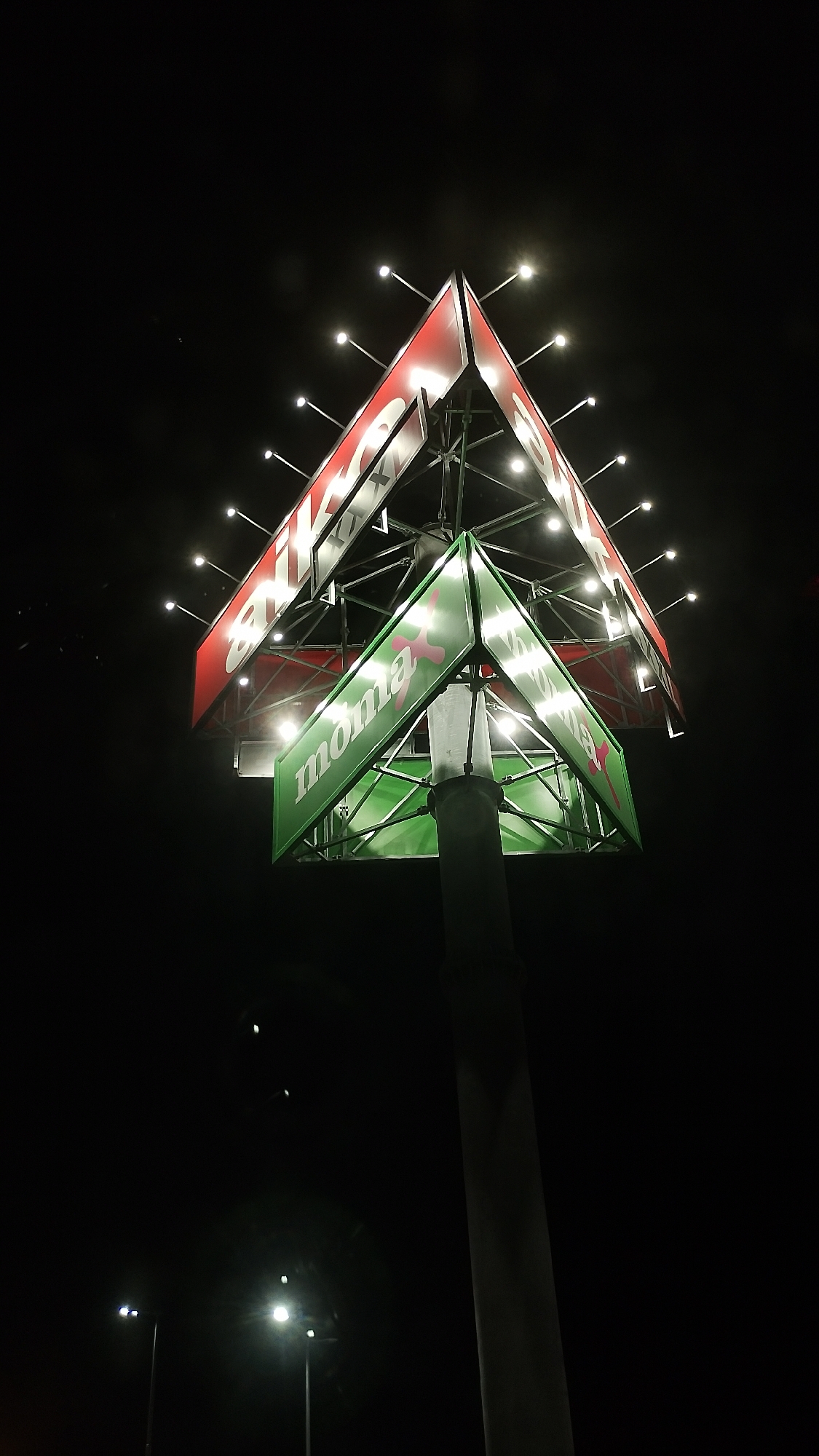 An illuminated billboard sign structure, triangular in shape and with multiple facets, is seen at night. The upper facets are red with white lettering that appears to say "DIKOB". The lower facets are green with white lettering that reads "mona". Each facet is outlined and illuminated by numerous small, bright white lights. The structure is supported by a thick concrete pole in the foreground. In the background, the night sky is dark, with several distant streetlights visible. The scene is shot from a low angle, looking up at the billboard. This billboard is located at the intersection of "Knyaz Aleksander I" Boulevard and "Hristo Botev" Street in Plovdiv, Bulgaria.