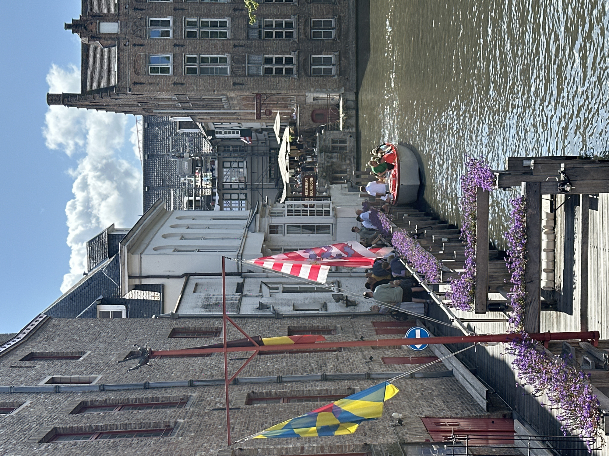 This is a photograph of a canal in Bruges, Belgium, likely taken on a sunny day. In the foreground, a wooden walkway lined with planters filled with purple flowers leads along the canal. A red pole stands near the walkway, from which two flags are flying: one with yellow, blue, and red geometric patterns, and another with red and white stripes and a crest. A blue directional arrow sign is visible on the pole.
On the walkway, several people are gathered, some standing and others appearing to be boarding or disembarking from a boat. The boat, which is red and white, is on the canal and carries a group of passengers. Across the canal, historic buildings with brick facades and various architectural details line the waterway. One building has an awning over an outdoor seating area, suggesting a restaurant or cafe. The canal water reflects the bright sky and surrounding buildings. The overall scene depicts a picturesque and bustling tourist area in Bruges.