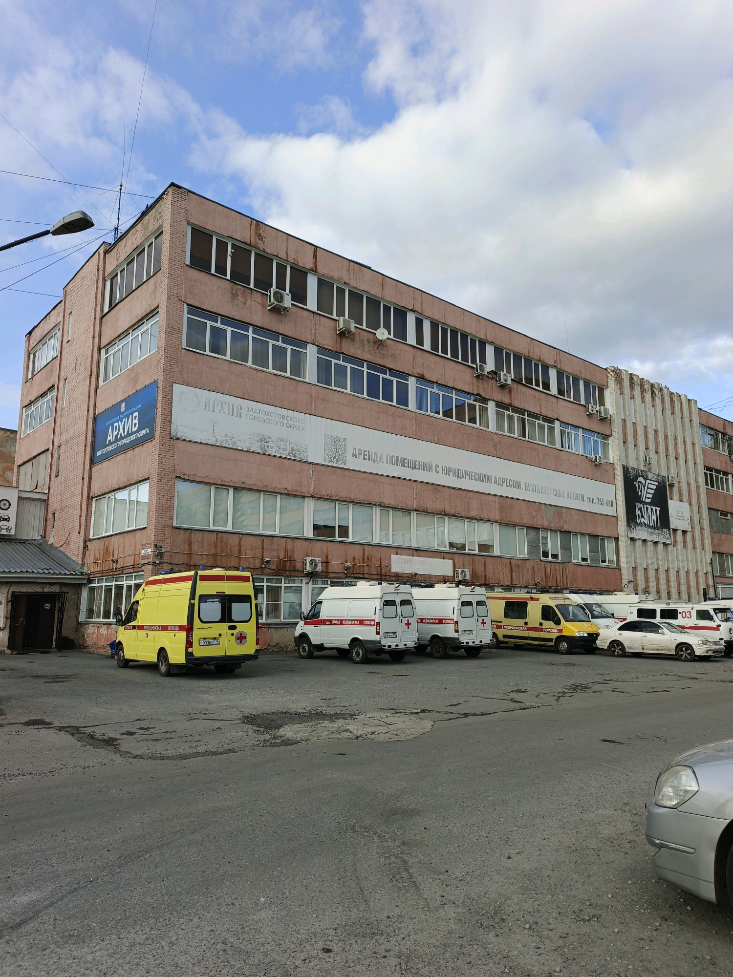 The image depicts an exterior view of a multi-story, pink brick building in Zlatoust, Russia. The building appears to be an office or administrative complex, with multiple windows on each floor. Several signs are visible on the building's facade. One prominent blue sign reads "АРХИВ" (Archive) with smaller text below indicating "ЗЛАТОУСТОВСКОГО ГОРОДСКОГО ОКРУГА" (Zlatoust Urban District). Another large banner features text in Russian and a QR code.
In front of the building, parked on an asphalt surface, are several vehicles. Most notably, there are at least three white vans with red crosses, suggesting they are ambulances or medical transport vehicles. One yellow ambulance is also parked prominently. A white sedan and another white van are also visible. The sky above is partly cloudy, with a mix of blue sky and white clouds. A street light pole extends into the frame from the left side of the image. The overall scene suggests a functional, urban environment, likely a business or government facility.