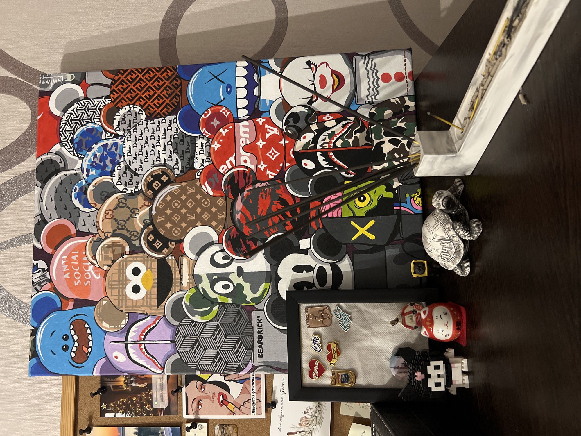 The image depicts a desk with various decorative items and artwork. A large canvas featuring a collage of stylized bear figures with different patterns, including brand logos like Supreme, Dior, Gucci, and Louis Vuitton, dominates the background. To the left of the canvas, a corkboard displays several postcards and photos, including a Christmas tree scene and a stylized portrait of a woman applying lipstick. Below the corkboard is a framed display of several pins, some depicting hearts with text, a deer, and a crest.
On the desk surface, in front of the framed pins, are several small figurines: a black and white pixelated character, a red and white round cat figurine (possibly a Daruma doll), and a small black cat with a yellow square on its head. A grey, detailed turtle figurine sits to the right of the cat figurine. Further to the right, a white incense holder contains several lit incense sticks, with ash scattered in the holder. The overall environment suggests a personal display space, likely in a home or office. There is no indication of the location being in Kazan, Russia.