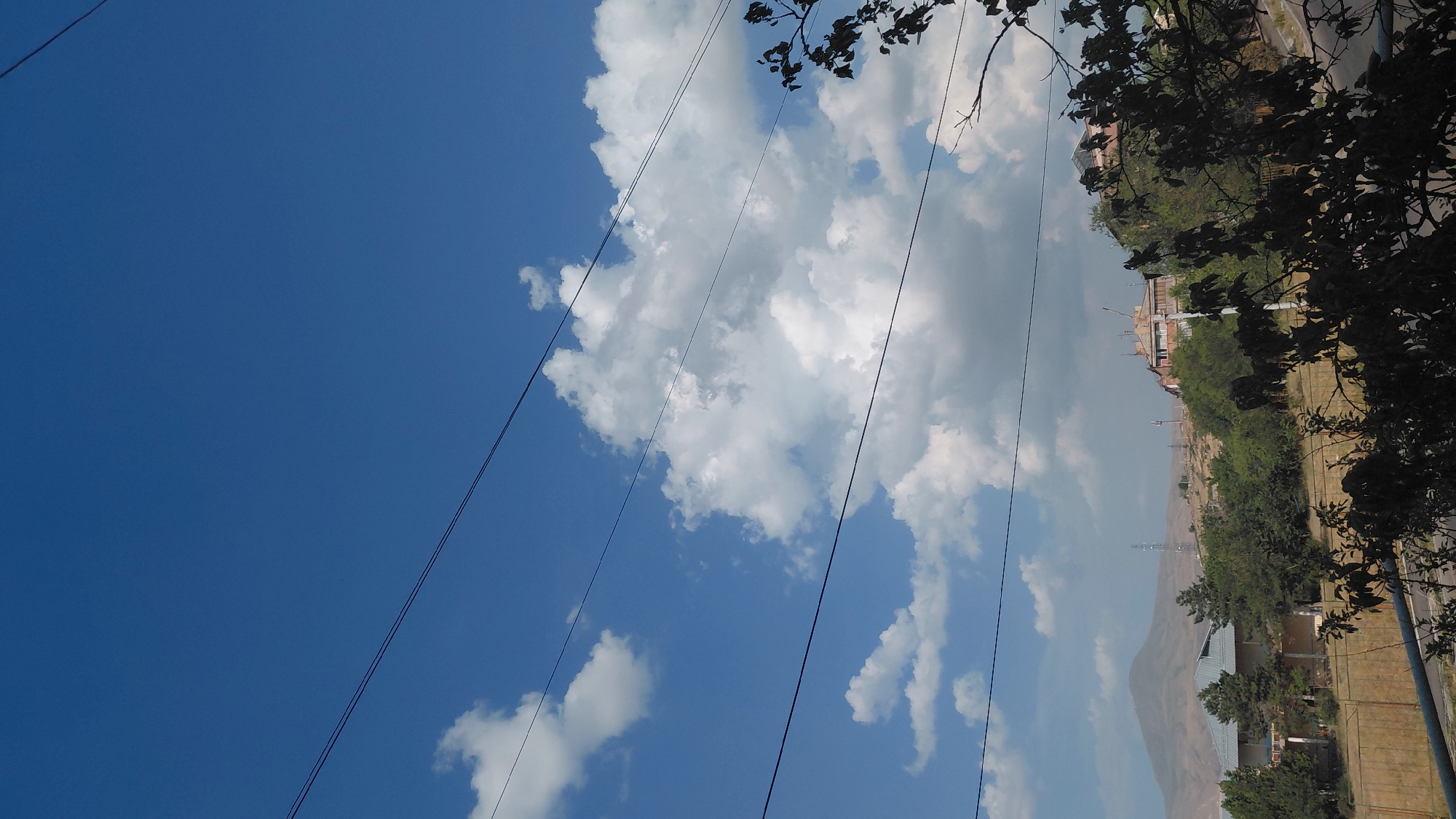 A landscape photograph taken from a slightly elevated viewpoint captures a clear blue sky filled with fluffy white clouds. Several dark power lines stretch horizontally across the upper portion of the frame. Below the sky, a hilly landscape unfolds. In the middle ground, several buildings are visible, nestled among lush green trees. One building, with a light-colored facade and multiple windows, appears to be a multi-story structure. Another building, with a terracotta-tiled roof and balconies, is situated on the higher slope. The distant background features a large, hazy mountain. In the foreground, dark silhouettes of tree branches and leaves obscure parts of the view. The image appears to be taken in Abovyan, Armenia, given the characteristic mountainous terrain and architectural styles. No people or discernable actions are visible.
