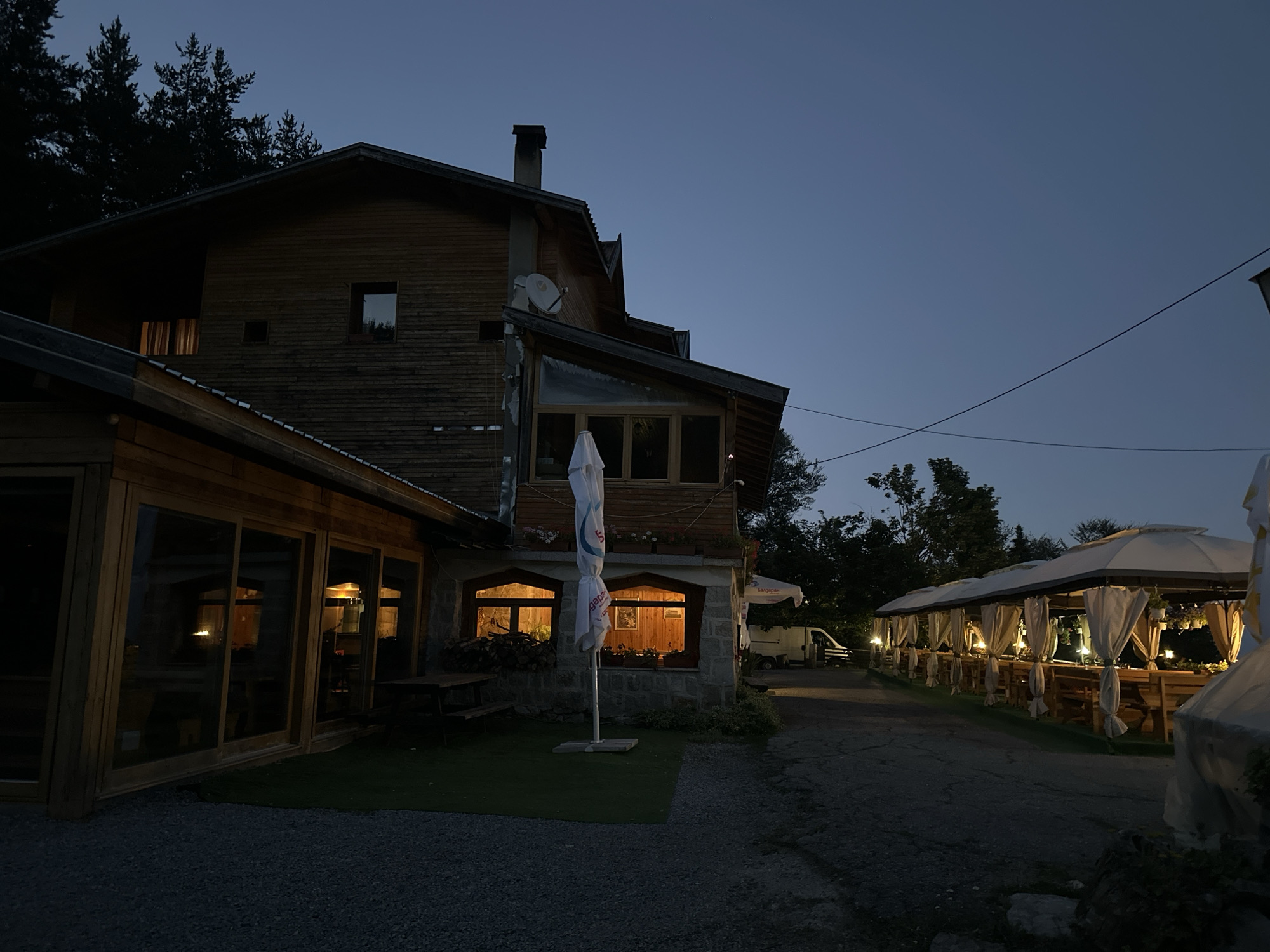 The image captures an exterior view of a wooden building with stone accents, possibly a restaurant or guesthouse, at dusk. The building is illuminated from within through several large windows, casting a warm glow. Outside, there are outdoor seating areas with umbrellas and white curtains, set up along a paved pathway. A white van is parked in the background. Trees and a dark blue sky complete the scene, suggesting a rural or semi-rural setting. The location is identified as Bogoslov, Bulgaria.