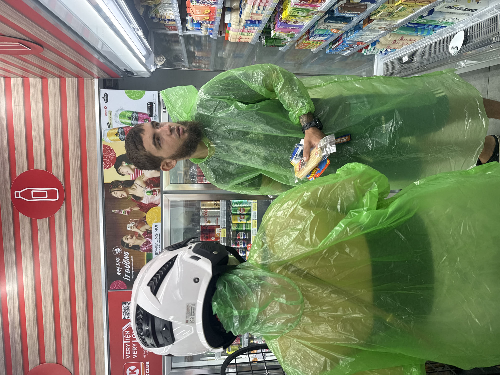 A man wearing a lime green disposable rain poncho and a white motorcycle helmet is standing in an aisle of a convenience store. He is looking to his right, towards a display of bottled drinks. In his left hand, he holds a small package containing a few toy cars and a snack. Partially visible to his left is another person wearing a similar lime green rain poncho, with a white helmet obscuring their head and neck. The store shelves behind the man are stocked with various beverages, including sodas and juice boxes. The store appears to be a 7-Eleven, based on the visible branding. The lighting suggests an indoor environment, likely within Nha Trang, Vietnam, a popular tourist destination.