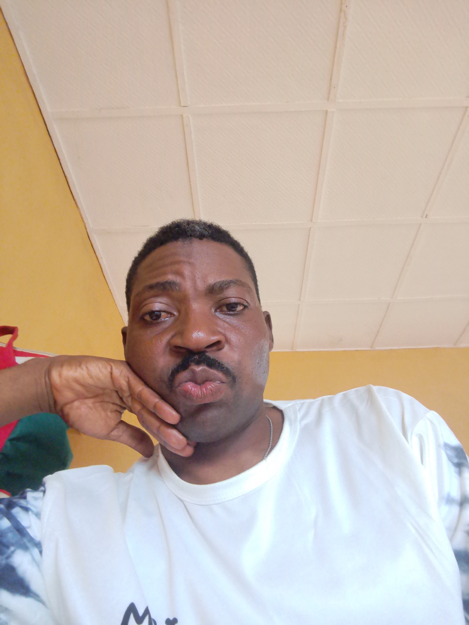 A medium-shot, eye-level selfie shows a Black man indoors, with his left hand resting on his chin and his mouth pursed. He is looking directly at the camera. He has a mustache and short, graying hair. He is wearing a white t-shirt with a tie-dye pattern on the sleeves and a silver chain necklace. The background consists of a yellow wall and a white, textured ceiling with square panels. This appears to be an indoor domestic setting, possibly a living room or bedroom in Ajasse Ipo, Nigeria.