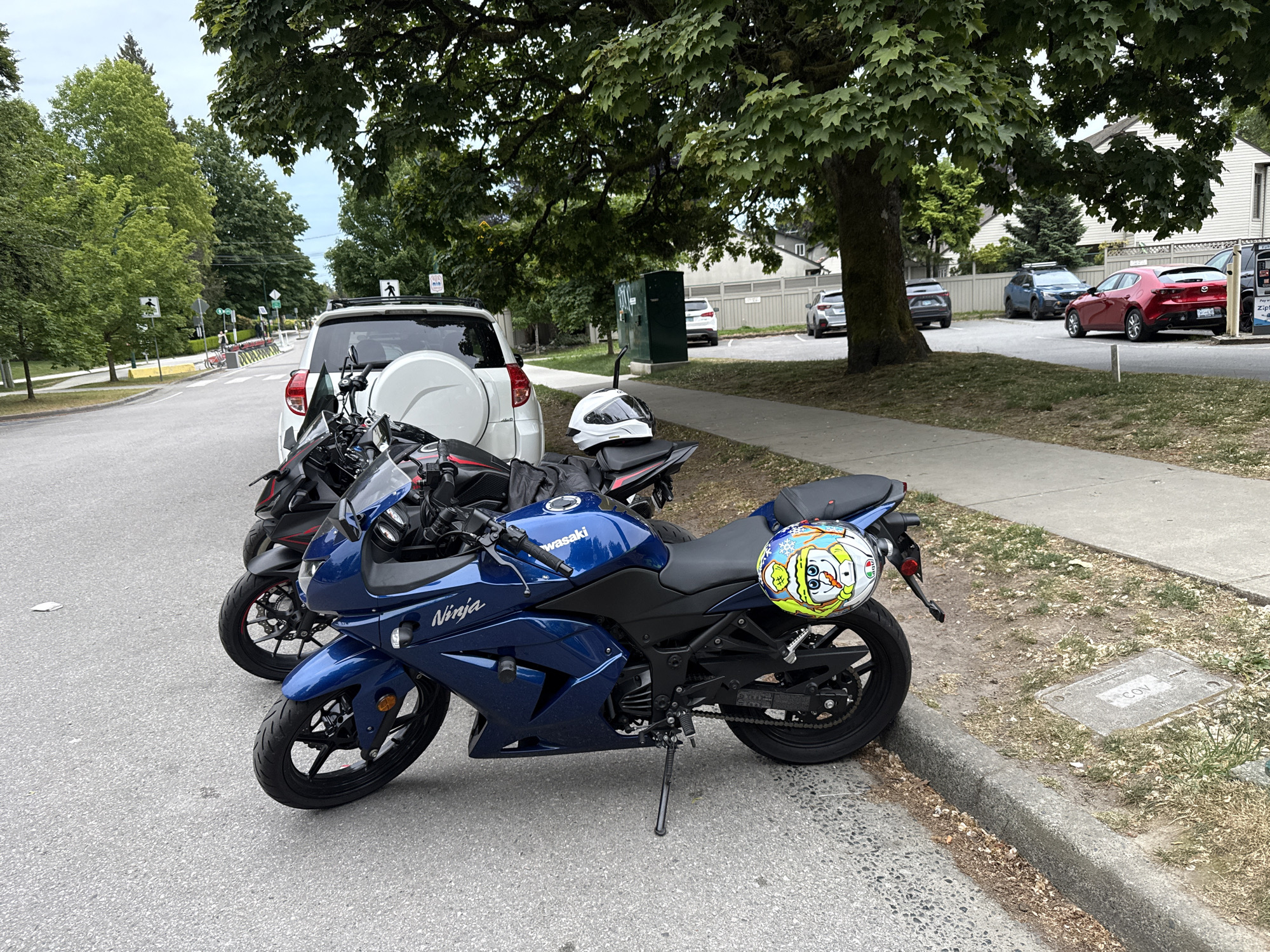 This photograph shows two motorcycles parked on the side of a road in Vancouver, Canada. The motorcycle in the foreground is a blue Kawasaki Ninja with a distinctive helmet featuring SpongeBob SquarePants as a snowman resting on the back. Behind it, partially obscured, is another motorcycle, appearing to be black, parked next to a white SUV with a rear-mounted spare tire. A white helmet sits on the black motorcycle's seat. A sidewalk runs alongside the road, with grass and a manhole cover visible between the sidewalk and the curb. Trees provide a canopy overhead, casting shadows on the scene. Several cars are parked further down the street. A pedestrian crossing sign is near the SUV. The location is likely in a residential or suburban area of Vancouver, given the presence of houses and parked vehicles.