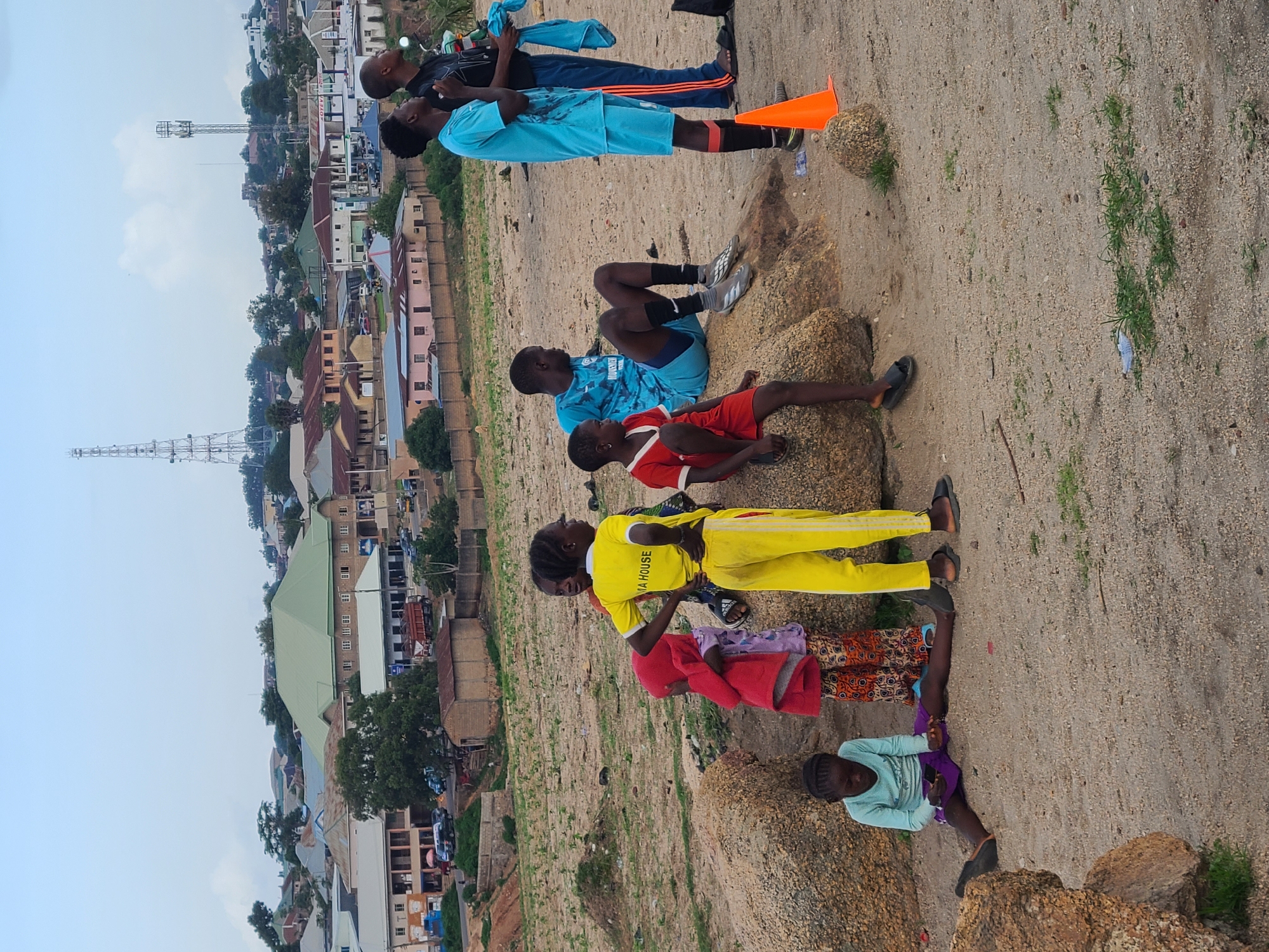 The image captures a group of children and young adults in the city of Jos, Nigeria, with a backdrop of residential buildings and telecommunication towers. In the foreground, several children are gathered on rocky terrain. A young girl in a light blue long-sleeved shirt and purple skirt sits on the ground, looking at a mobile phone. To her right, a girl in a bright yellow shirt and yellow pants stands with her hands on her hips. Next to her, a child in a red hooded jacket and patterned pants stands partially obscured.
Further back, a young man in a light blue soccer jersey and shorts sits on a rock, looking to the right. Another young man in a similar light blue soccer outfit stands facing away from the camera, holding a blue towel. He is wearing a black undershirt and has black stripes on his pants. A child in a red shirt and dark shorts is seated on a rock to the left of the young man in the blue jersey. An orange traffic cone is visible on the sandy ground to the right of the standing young men.
The environment is a mix of dry, sandy ground with scattered rocks and patches of green grass. In the middle ground, a collection of buildings with various roof colors, including green, beige, and pink, are visible. Several vehicles, including cars and a truck, are present on a road winding through the settlement. Two prominent telecommunication towers stand tall in the background, silhouetted against a pale sky. The overall scene suggests an informal gathering in an urban or semi-urban area of Jos.