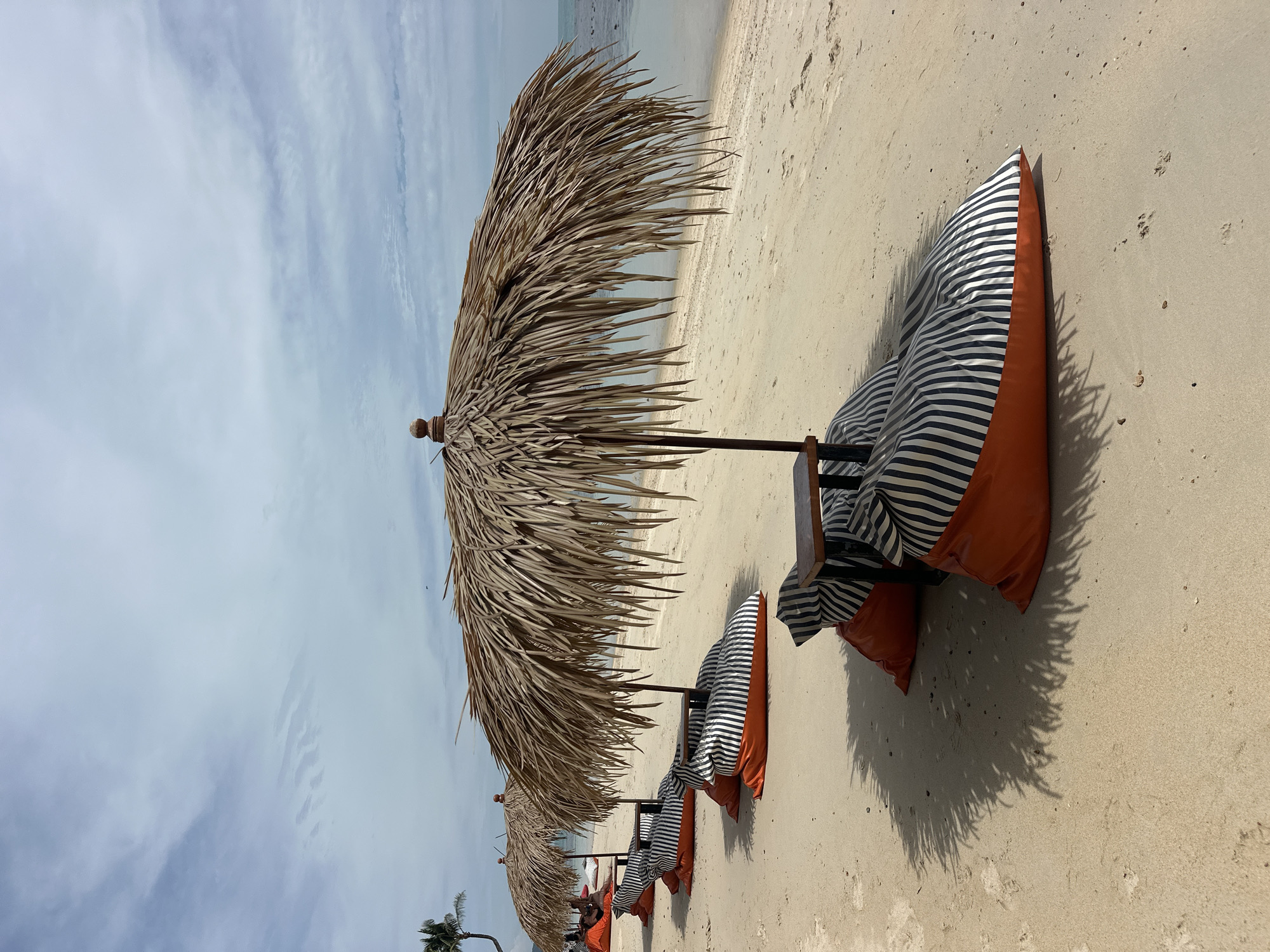 On a sandy beach in Koh Samui District, Thailand, a row of beach chairs, each consisting of an orange beanbag base with a striped cushion and a small wooden table, are arranged under thatched umbrellas. The umbrellas provide shade on the light-colored sand, which is bordered by the calm, blue ocean. The sky is partly cloudy with a few wisps of white clouds against a pale blue backdrop. A lone palm tree is visible in the distance on the left side of the frame, suggesting a tropical setting. The overall scene depicts a relaxed and inviting beachside lounging area.