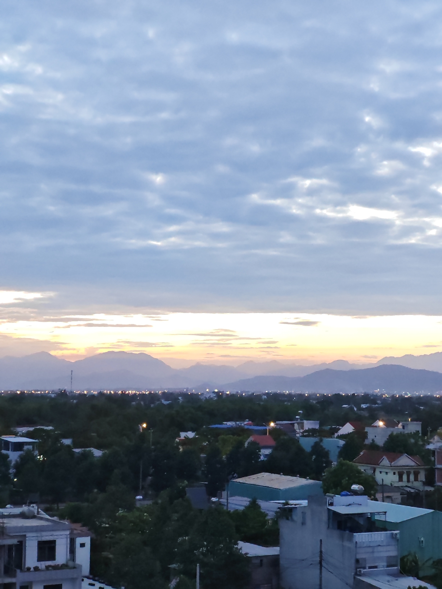 The image captures a view of the city of Đà Nẵng, Vietnam, at dusk. The sky is filled with layered clouds in shades of blue and grey, with streaks of orange and yellow near the horizon, indicating the setting sun. In the distance, a range of hazy mountains forms a silhouette against the sky. Below the mountains, a city lights twinkle, suggesting a developed urban area. The foreground features a dense canopy of dark green trees interspersed with rooftops of various buildings. Several structures are visible, including houses with red and white roofs, and some with flat, light-colored roofs. One building in the lower left appears to be a multi-story residential building with balconies. The overall scene suggests a suburban or semi-urban environment within Đà Nẵng.