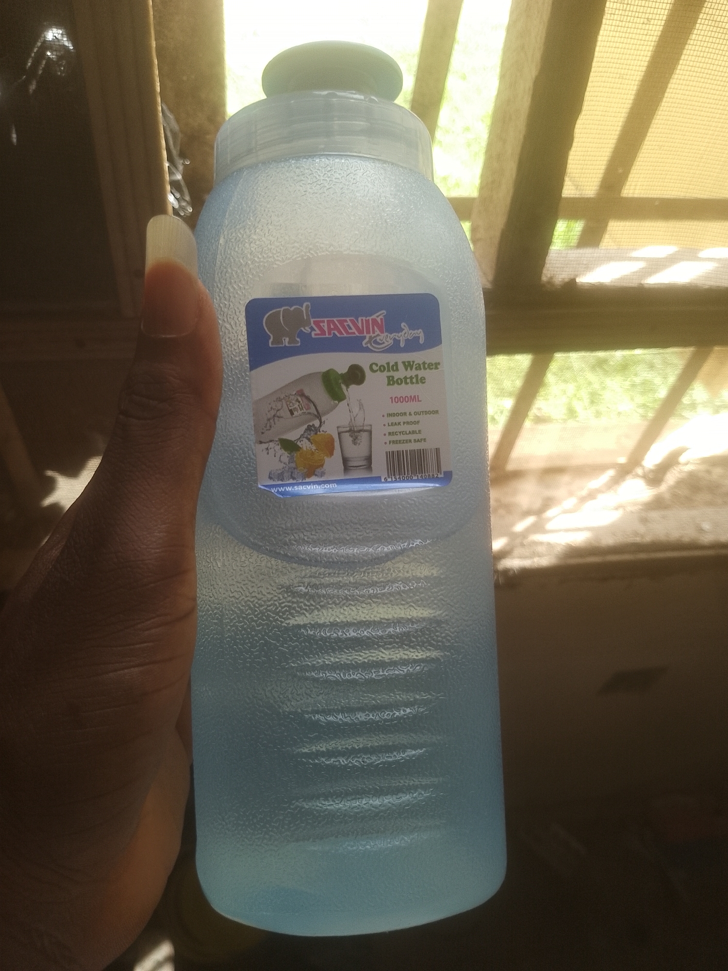 A light blue, textured plastic water bottle is held in a person's hand. The bottle has a blue and white label with text and an illustration of water pouring into a glass, along with lemons. The label reads "SACVIN Cold Water Bottle 1000ML INDOOR & OUTDOOR LEAK PROOF RECYCLABLE FREEZER SAFE". The background shows a wooden structure with netting and foliage, suggesting an outdoor or semi-outdoor setting. The lighting suggests it is daytime. The scene is not specifically identifiable as being in Majemu, Nigeria; it is a generic image of a water bottle.