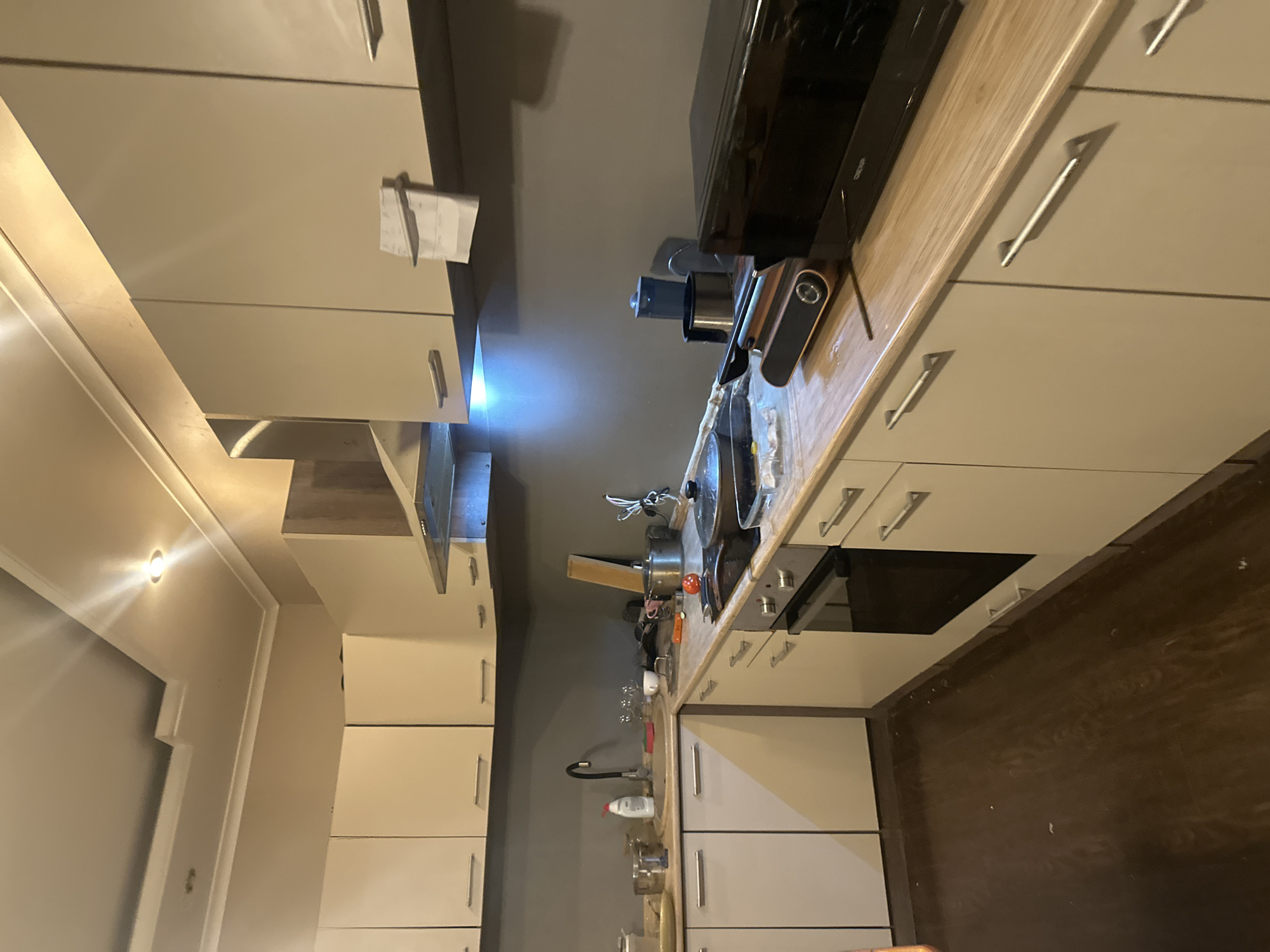 The image depicts a kitchen interior. The kitchen features light-colored cabinetry, a dark countertop, and a dark wood floor. The room appears to be in a state of disarray, with various kitchen items scattered on the countertops and sink area, including pots, pans, dishes, and a bottle of dish soap. There is a faucet over the sink, a stovetop with cookware, and a microwave oven. A ventilation hood is visible above the stovetop. The lighting in the room is a mix of overhead lights and under-cabinet lighting, casting shadows and highlighting certain areas. The overall impression is one of a lived-in and possibly recently used kitchen. The location is identified as Ореховка, Russia, however, there is no visual information in the image to corroborate this specific geographical location.