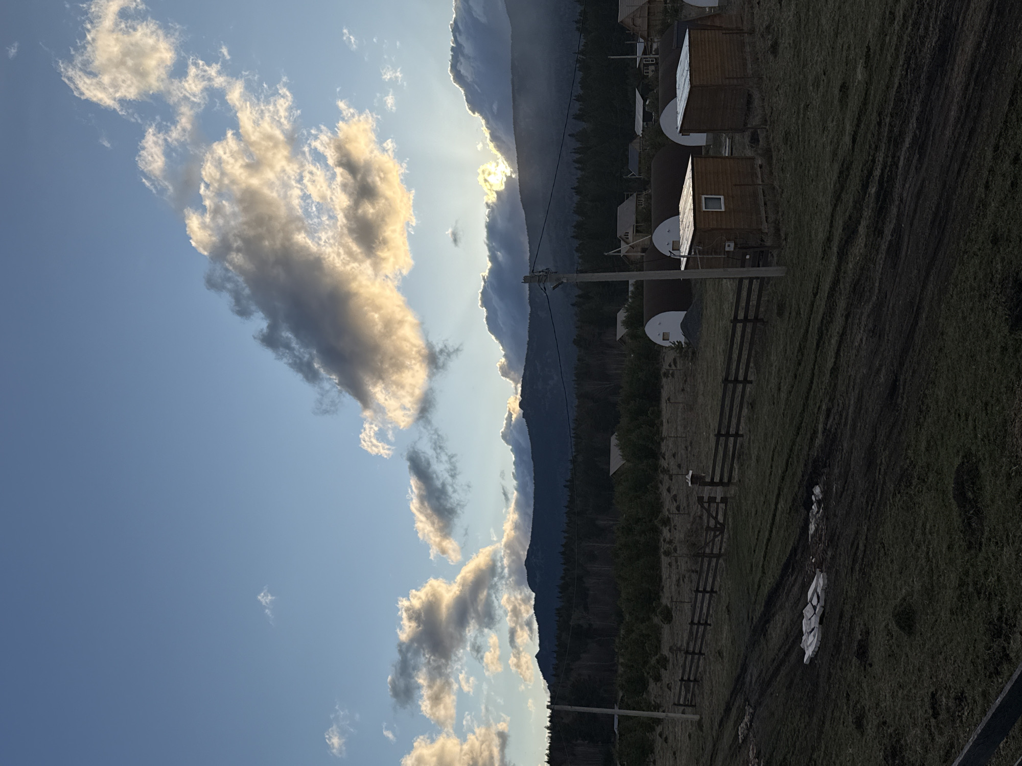 The image depicts a landscape with several features. In the background, there's a mountain range, partly obscured by clouds. The sun's rays peek through the clouds near the mountain. In the mid-ground, there are wooden structures resembling cabins or sheds with white-domed annexes, potentially storage or utility units. A wooden fence runs through the scene, and utility poles with wires are visible. The foreground consists of grassy terrain with dark, possibly muddy paths. The overall lighting suggests either sunrise or sunset. The location is Tyulyuk, Russia, judging by the surrounding environment and architecture. There are no people visible in the image.