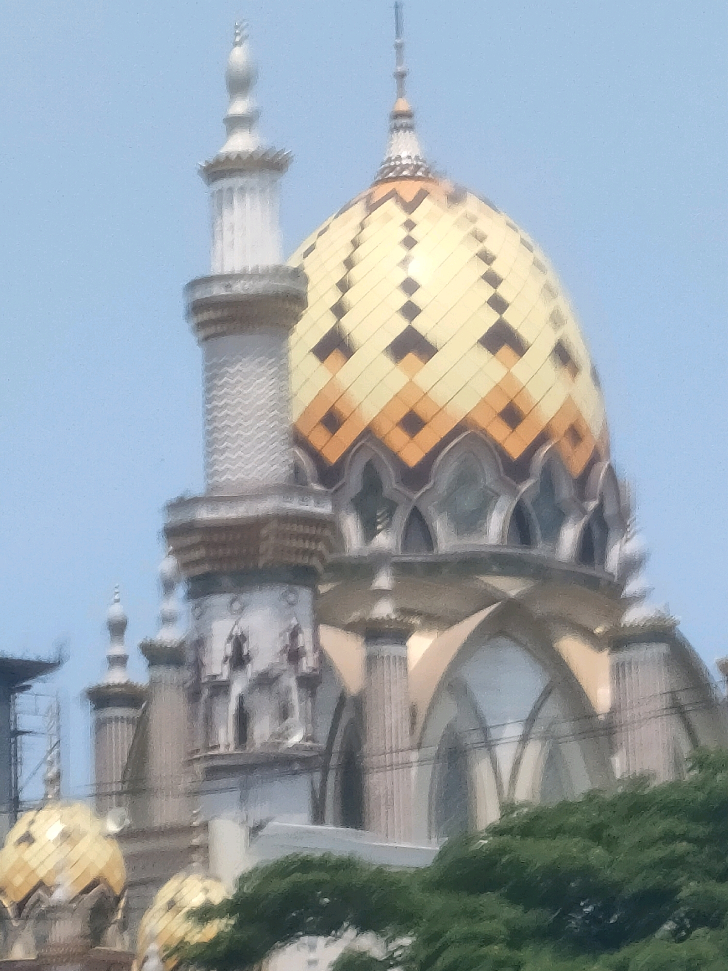 The image shows a close-up view of the Great Mosque of Surabaya, located in Surabaya, Indonesia. The mosque features a prominent golden dome adorned with an intricate geometric pattern of yellow and dark orange tiles. To the left of the dome, a tall, slender minaret with a silver, tiered spire rises against a clear blue sky. The minaret is decorated with vertical fluting and a textured pattern. The main structure of the mosque exhibits classical architectural elements, including arched windows and ornate columns. In the foreground, lush green trees obscure the lower portion of the building, partially concealing smaller golden domes. The overall impression is of a grand and visually striking religious edifice.