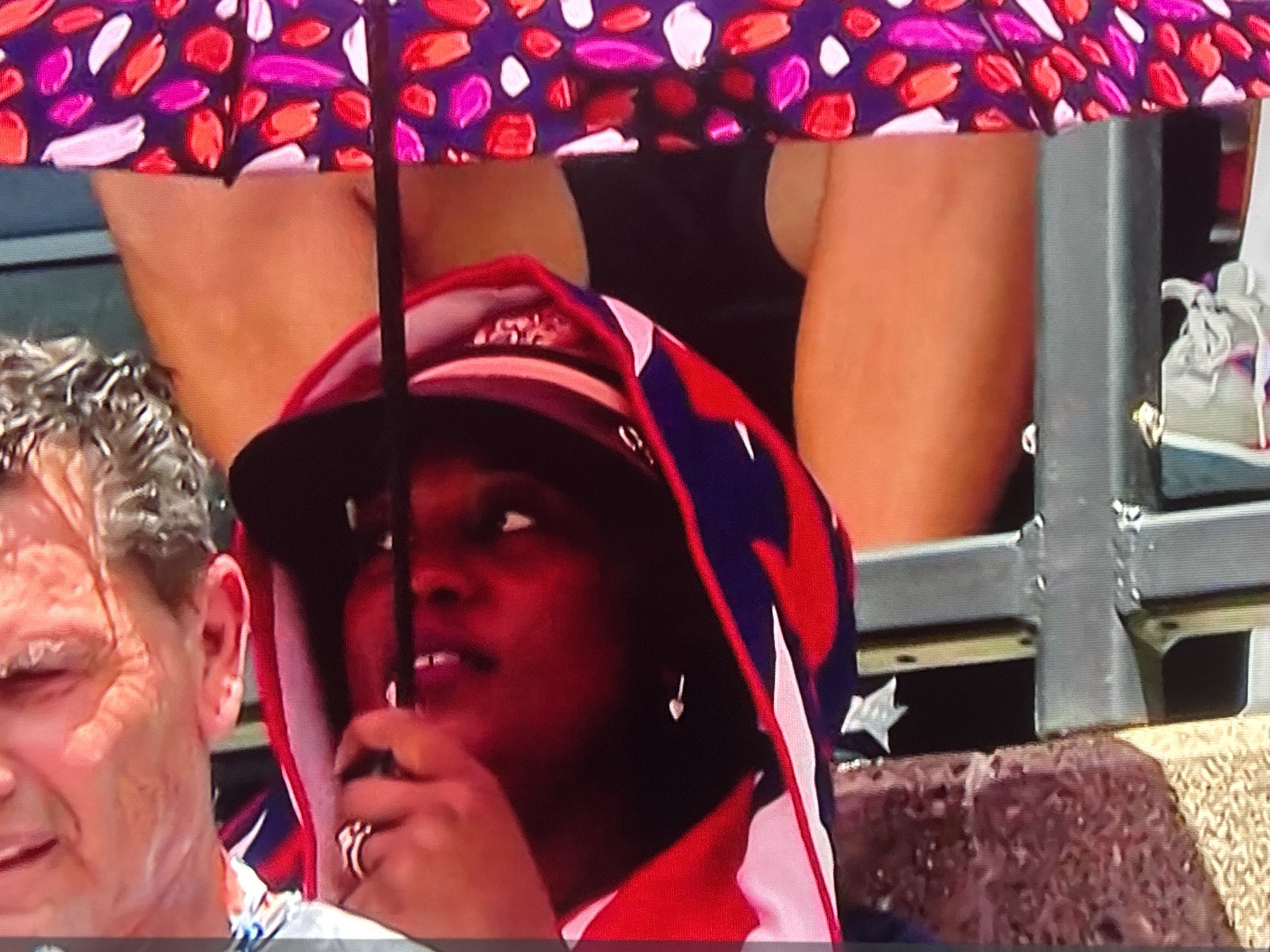 The image is a close-up, slightly blurry, and overexposed photograph of a woman wearing a hooded jacket and holding a patterned umbrella. Her face is visible, and she is looking off to the side with her mouth slightly open. She is wearing a ring on her finger and a small heart-shaped earring. To her left, a man's face is visible, and he appears to be crying or sweating profusely. The background shows a blurry view of spectators, a metal railing, and a shoe. The lighting is harsh, casting red tones on the subjects. The overall setting appears to be an outdoor event, possibly a sporting match.