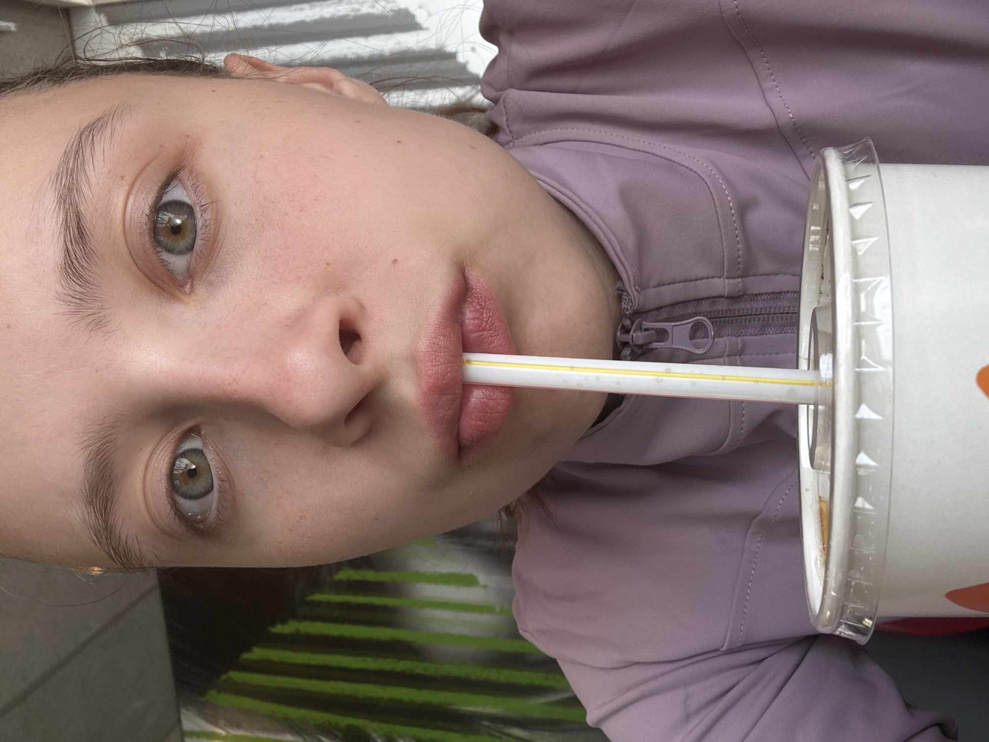 A young woman with light skin and light-colored eyes is drinking from a white cup with a yellow straw. She is wearing a light purple jacket with a zipper visible. The background includes what appears to be a table with a green and black design. It's not possible to determine if the location is Perm, Russia without additional context within the image.