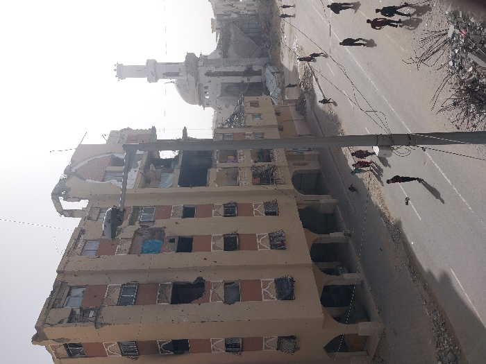 A severely damaged building stands tall, bearing multiple holes and structural damage. A mosque with a prominent dome and minaret is visible in the background, also appearing damaged. A wide road runs parallel to the building, with several pedestrians walking along it. Debris litters the side of the road. The overall scene conveys destruction and displacement.
