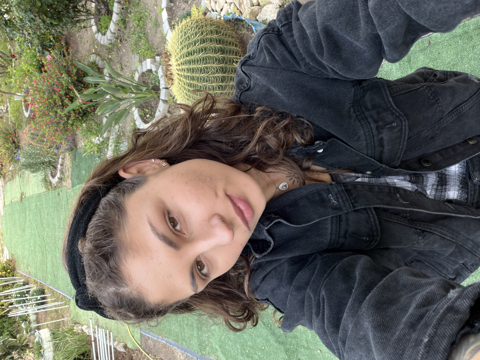 A person is taking a selfie outdoors in Kiryat Bialik, Israel. The individual has brown hair and is wearing a black denim jacket over a black and white plaid shirt. They have a nose ring and multiple earrings. Behind the person, there's a cactus in a pot surrounded by rocks and various other plants and greenery. There is also a patch of artificial green turf. In the background, partially obscured, there are metal supports.