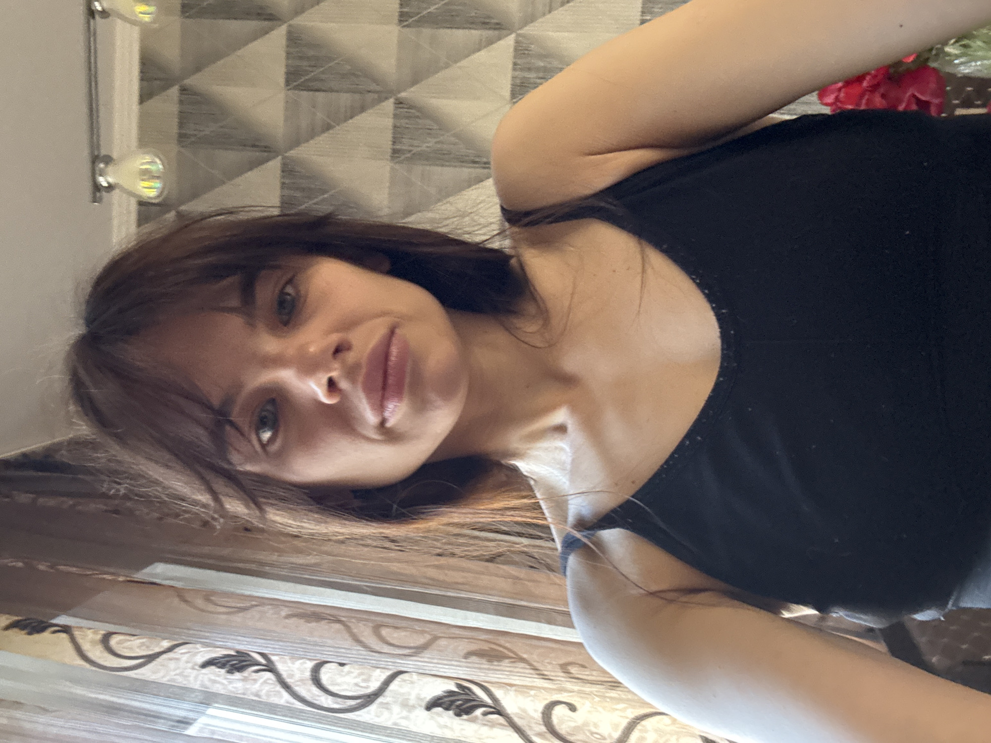 This indoor portrait features a young woman, likely taken in Rostov-on-Don, Russia. The image captures her in a relaxed pose, possibly a selfie, looking directly at the camera with a soft, faint smile. She has long, dark brown hair, with some strands falling across her forehead and shoulders, and light-colored eyes. She is wearing a plain black tank top or camisole.
The background reveals elements of a modern interior. Behind her head, a wall is decorated with geometric patterned wallpaper, displaying a repeating motif of triangles or diamond shapes in light and dark grey or beige tones. Two spherical light fixtures with iridescent bulbs are mounted on this wall to her left. Below the wallpaper, a patterned curtain or set of blinds with ornate, swirling designs in brown and beige hues adds a decorative touch. To her right, a vase containing several red flowers, possibly artificial, is visible. The lighting is soft and even, highlighting her features. No visible text is present in the image.