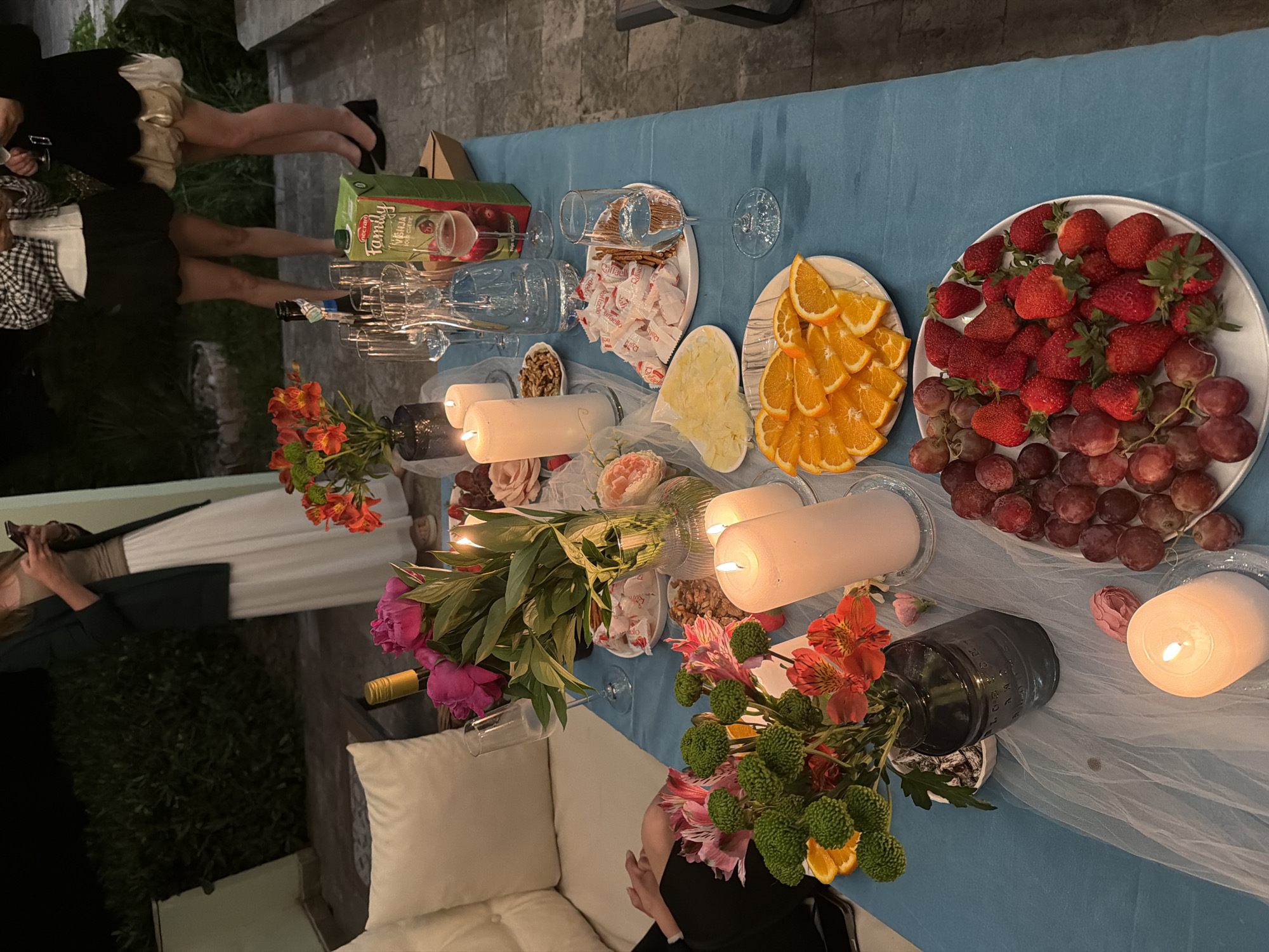 The image shows a table set for an outdoor gathering. The table is covered with a blue cloth and decorated with lit candles, flowers (including roses, asters, and orange flowers), and a sheer white runner. Food items include strawberries, grapes, sliced oranges, cheese, and wrapped candies. Wine glasses and a juice box are also present. People are present around the table, with legs visible near the juice box and someone seated near a white cushion. The location appears to be outdoors, possibly a patio or garden. The scene is not definitively identifiable as Šušanj, Montenegro, due to a lack of specific identifiable local landmarks.