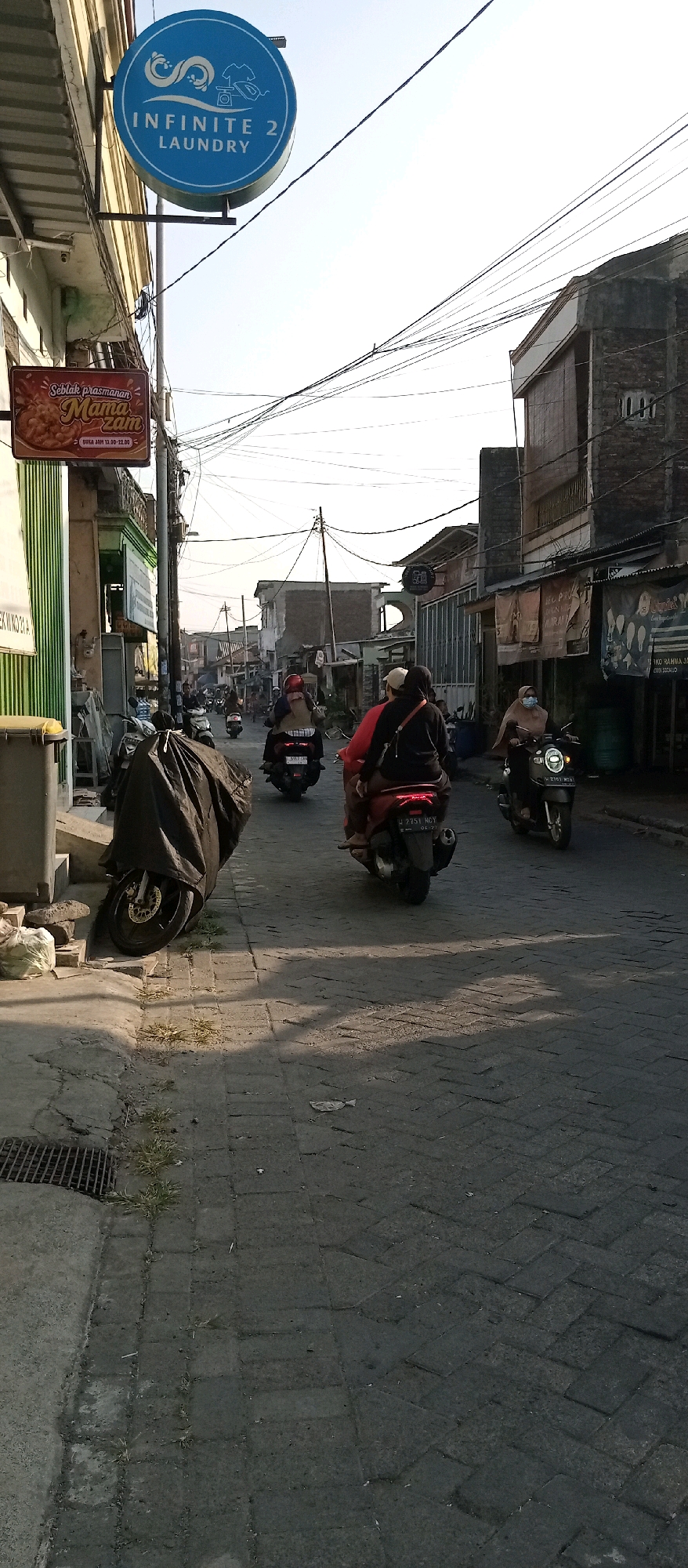 In Wadungasri, Indonesia, a street scene unfolds with multiple motorcycles and riders navigating a paved road. A prominent blue circular sign with white text reads "INFINITE 2 LAUNDRY" and is positioned above a smaller rectangular sign advertising "Seblak prasanan Mama Zam". On the left side of the image, a motorcycle is covered with a black tarp. The street is lined with buildings and overhead electrical wires are visible against a bright sky. Several individuals are riding motorcycles, some wearing face masks. The road surface is made of interlocking brick pavers. Shadows are cast across the road, indicating the time of day is likely late afternoon or early morning.