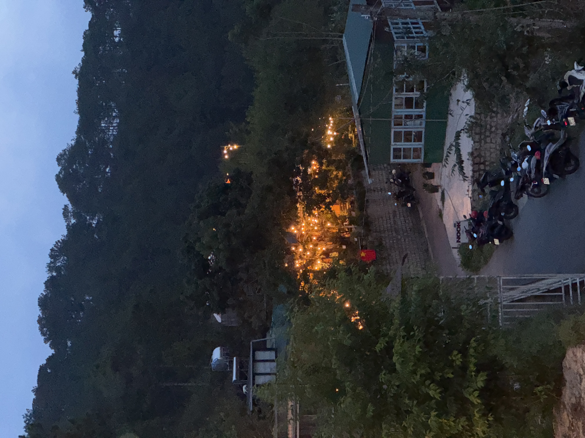 The image shows an elevated view of Xuân Trường, Đà Lạt, Vietnam at dusk. A dark, forested mountain range borders the left side of the frame, contrasting with the fading blue sky. A structure with a green roof and several windows stands in the middle right. A row of parked motorcycles is present. String lights illuminate a pathway. A Vietnamese flag is visible. A sign with the word "NOOK" is present. Various trees and shrubs fill the lower portion of the frame.