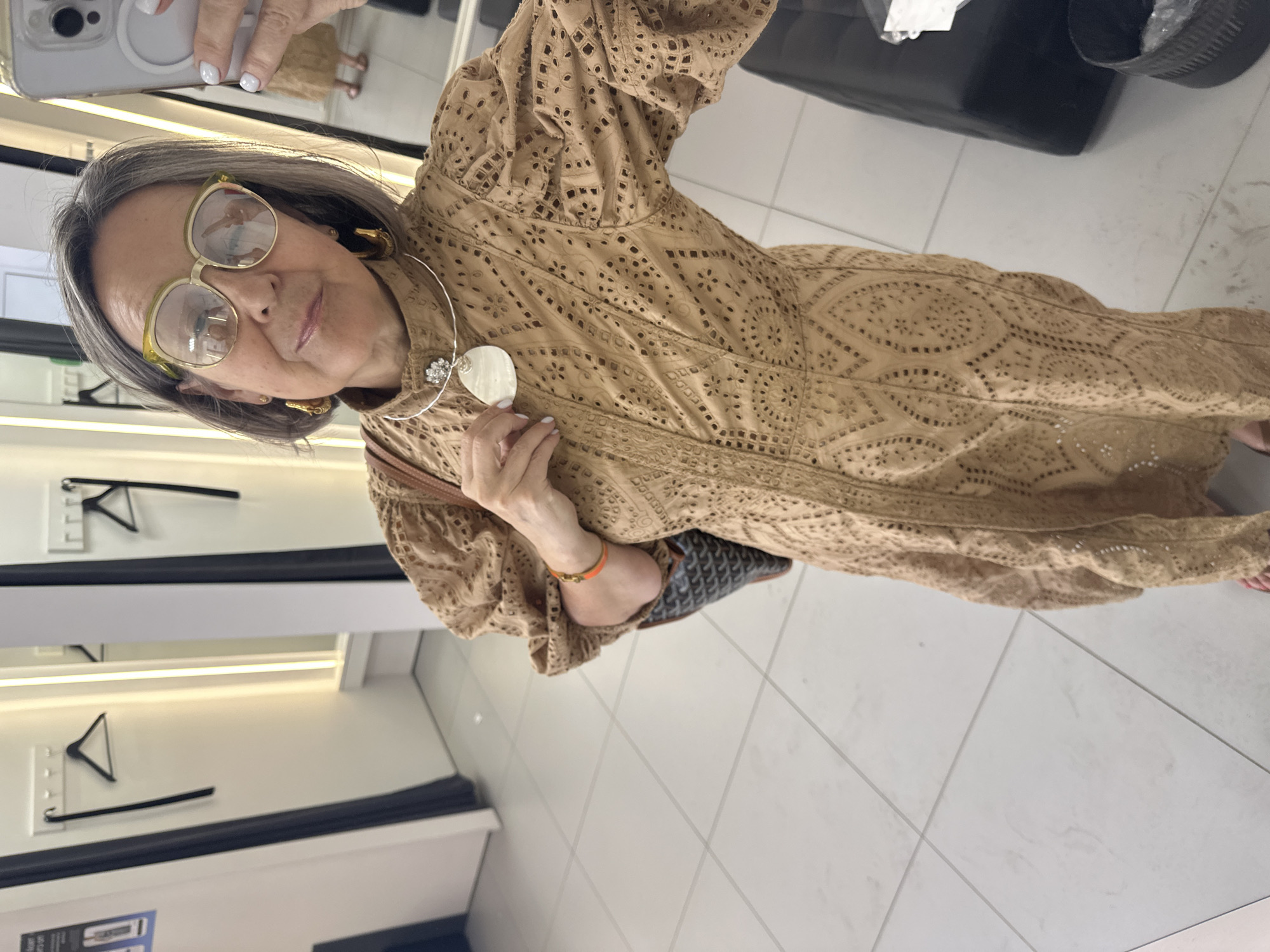 A woman with short gray hair and glasses is taking a mirror selfie in a brightly lit fitting room. She is wearing a tan, intricately patterned dress with puffed sleeves and is accessorized with large gold hoop earrings, a statement necklace, and an orange bracelet. A black and brown patterned tote bag is slung over her shoulder. The fitting room has white tiled floors and white walls with mirrors. A rack of clothes is visible in the background.