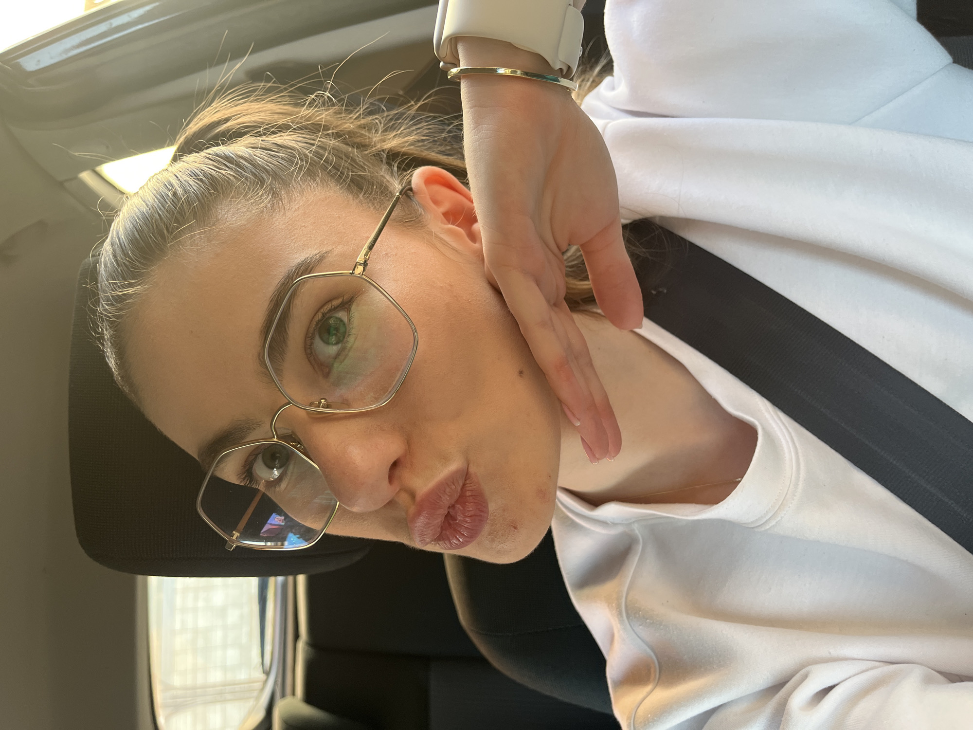 A fair-skinned woman with brown hair pulled back in a ponytail is posing for a selfie in what appears to be a car. She is wearing gold-framed glasses and a white sweatshirt. A black seatbelt crosses her chest. She has her right hand near her face, wearing a white smartwatch on her wrist and a gold bracelet. The headrest of the car seat is visible behind her head. Outside the car window, a tall building can be seen. The location of the car and the building is Unknown, Unknown.