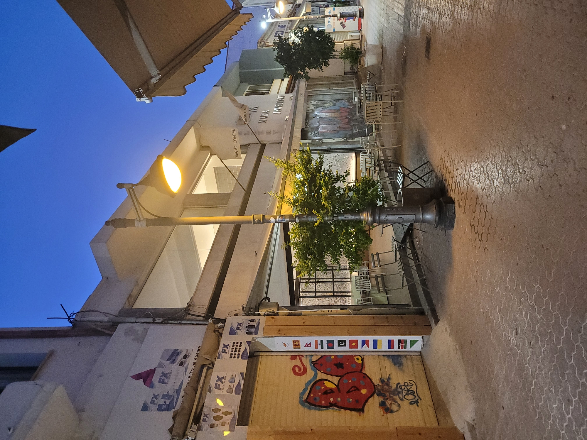The image depicts a street scene in Limassol, Cyprus, during twilight. The street is paved with textured interlocking blocks. On the left, a building features a wooden-shuttered shopfront with graffiti. Above the shutter, signage for "FX Accessories" and "FX for & Travel Agency" is visible. A street lamp with a lit bulb stands in the foreground, illuminating the area. To the right, another building houses establishments including "WAGMI COFFEE" and "MARIA JEWELLERY". Outdoor seating with chairs and tables is arranged in front of these businesses, some under awnings. The sky is a deep blue, indicating the transition from day to night.