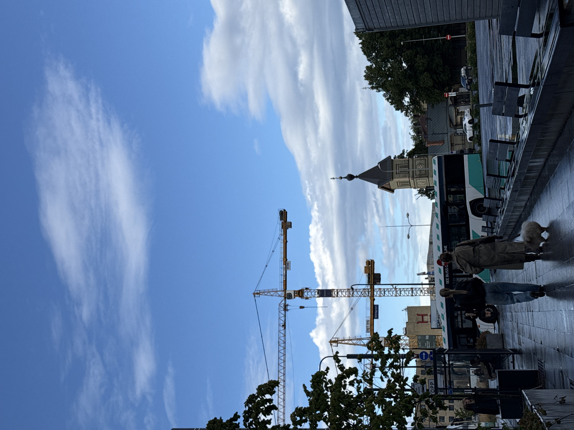 The image captures a street scene in Tallinn, Estonia, with construction cranes dominating the skyline. In the foreground, two women walk along a wet pavement, one with a small, fluffy white dog. They are walking away from the camera, towards a bus that is stopped at a bus stop. The bus is white and green, with text that reads "SEE BUSS G LINE". The pavement is slick with water, suggesting recent rain.
Further down the street, several tall construction cranes are visible, indicating ongoing development. Behind them, a distinctive clock tower with a dark spire stands out against the blue sky, characteristic of Tallinn's architecture. Trees with green foliage line the street, and various buildings, including a modern one with a large 'H' logo, are present. In the background, cars are parked and driving on the road, and streetlights can be seen. The sky is partly cloudy with patches of blue. The overall atmosphere is that of a city undergoing construction, with everyday urban life continuing amidst the development.