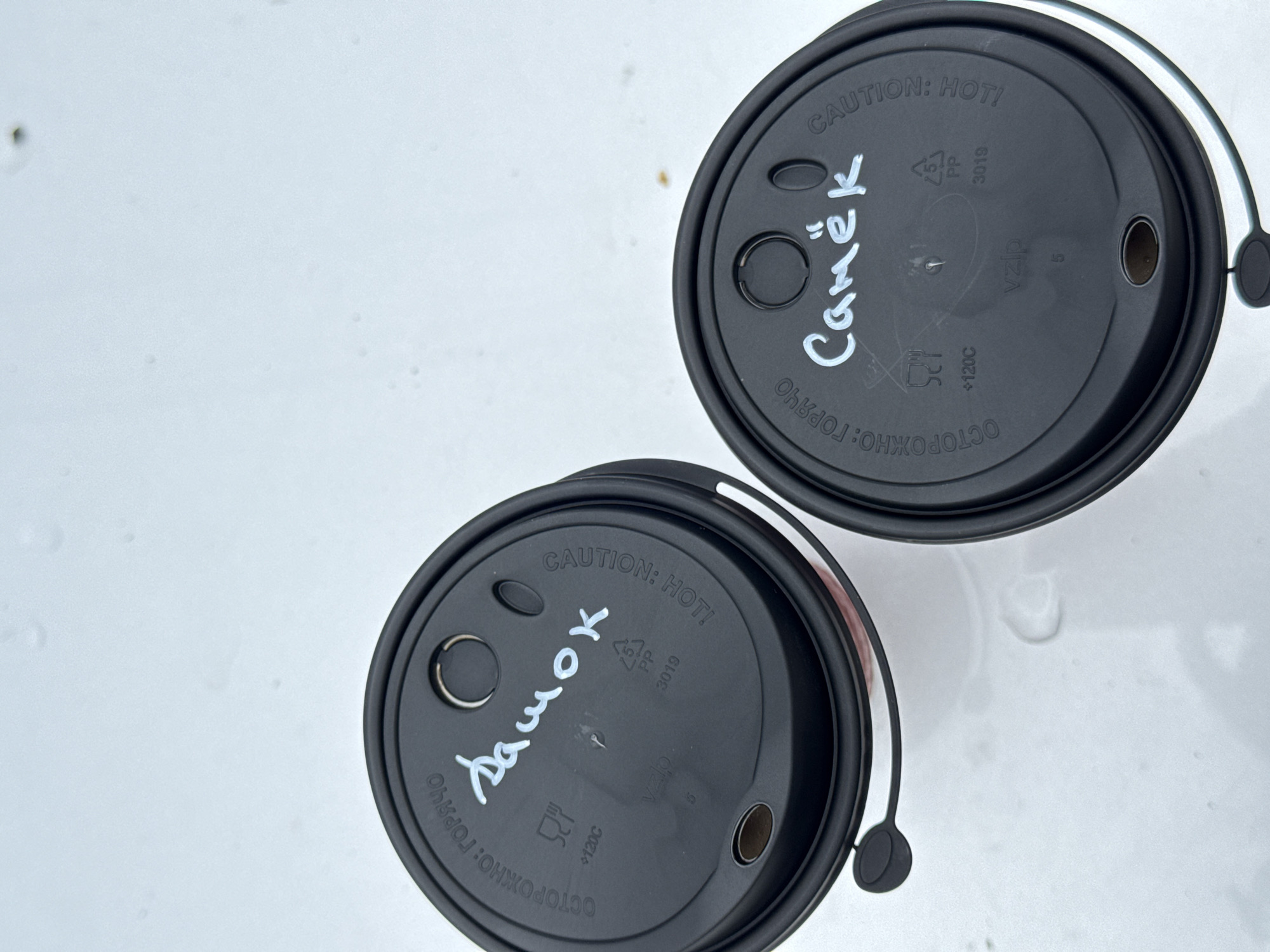 The image shows two black disposable coffee cup lids on a light-colored, possibly wet, surface. Both lids have text printed on them. The lid on the left has the Russian words "ОСТОРОЖНО: ГОРЯЧО" (Caution: Hot) printed in a curve around the edge. In the center, written in white marker, is the word "Дашок". There are also some symbols and "120C" printed on the lid. The lid on the right also has "ОСТОРОЖНО: ГОРЯЧО" printed around the edge and "CAUTION: HOT!" printed in a curve. In the center of this lid, written in white marker, is the word "Самёк". Similar symbols and "120C" are present on this lid as well. The surface they are resting on appears to be outdoors, possibly a street or sidewalk, given the slight wetness and light reflections. The location is not specifically identifiable as Chaikovsky, Russia based on the visual evidence.