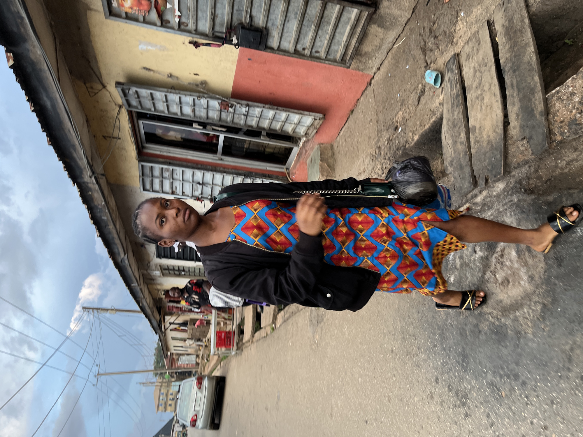 A young woman stands on an urban street, looking directly towards the camera. She wears a vibrant, short-sleeved dress with a geometric pattern in shades of blue, orange, red, and yellow, layered with a black bomber jacket. Her feet are adorned with black sandals featuring gold straps, and she carries a black bag over her left shoulder. Her hair is styled in braids.
The street is paved with asphalt or concrete, showing some cracks and wear. To the woman's right, lining the street, are buildings that appear to be shops or residences. These structures feature closed corrugated metal security shutters and walls painted in muted yellow and reddish-orange tones. A makeshift path or covering consisting of two wooden planks is visible on the ground beside the buildings, with a small blue plastic cup resting nearby.
In the background, the street continues, revealing a silver or white sedan parked further away and blurred figures of other people. Overhead, utility poles and a network of wires are visible against a partly cloudy sky. The scene depicts a casual, daytime moment in what appears to be an active street environment in Jos, Nigeria. No legible text is discernible in the image.