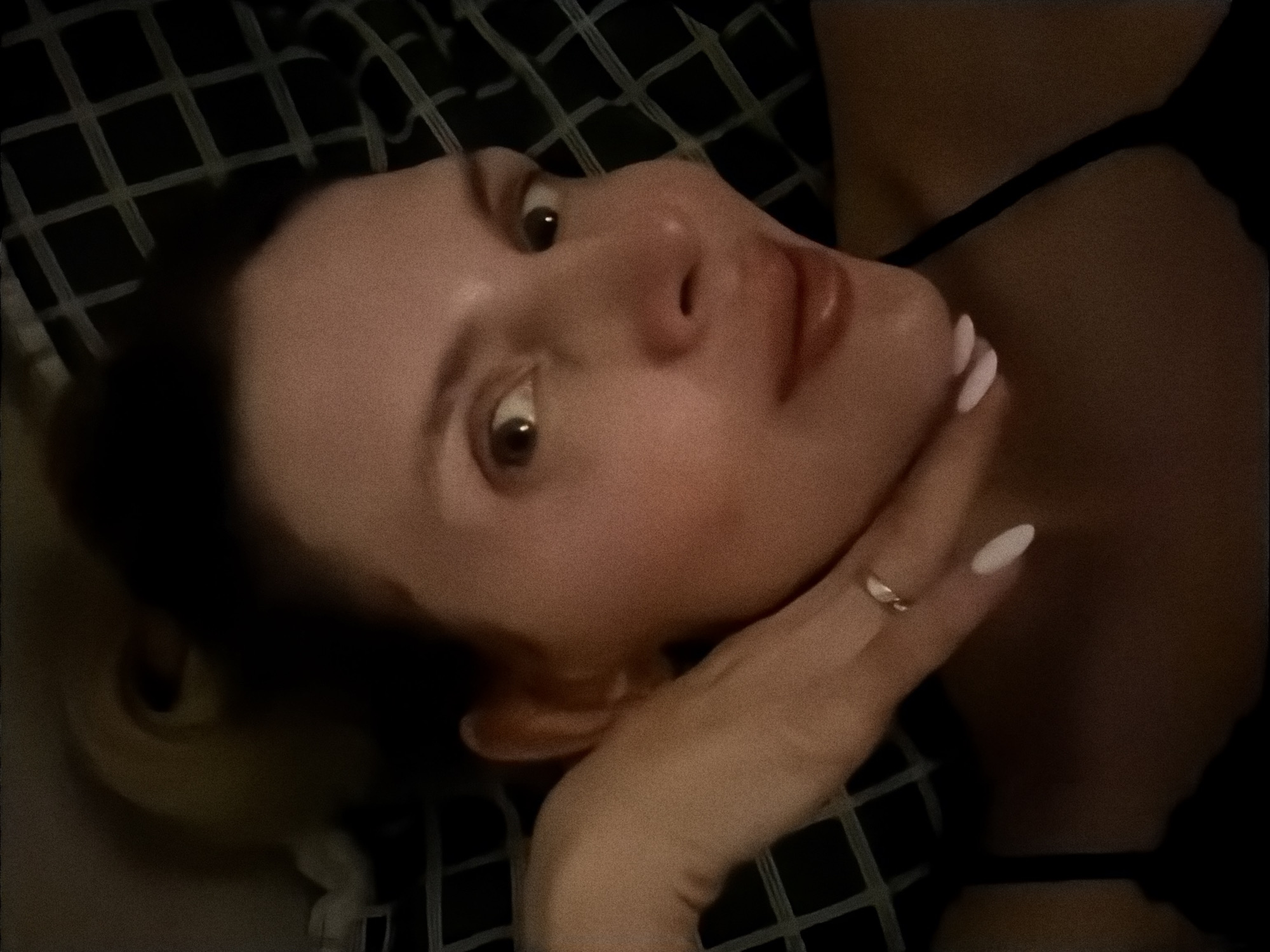 A woman is lying on a checkered blanket, looking up at the camera. She is resting her chin on her hand, which has a ring on one finger and white nail polish. She is wearing a thin-strapped top. The location is unknown and cannot be determined from the image.