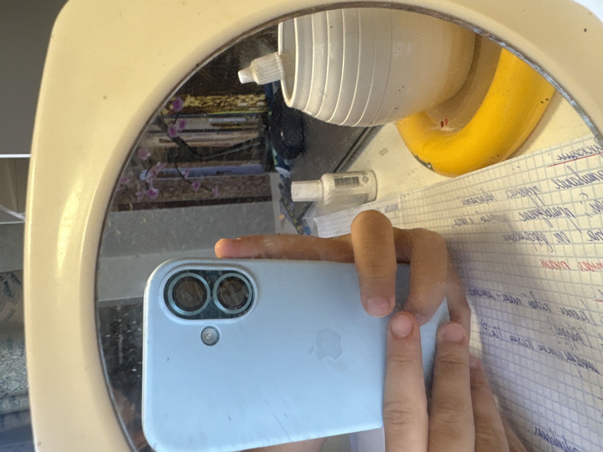 The image is a reflection in a mirror. In the foreground, a person's hands are holding a light blue smartphone with a dual-camera system. The smartphone has an Apple logo embossed on its back. The reflection shows a desk with various items, including a white ceramic vase with a white dropper inside, a small white bottle, and a yellow circular object. In the background of the reflection, a bookshelf with books and some pink flowers can be seen. The overall environment appears to be an indoor setting, likely a room with a desk and a mirror. There is no indication of the location being in Georgiyevsk, Russia.