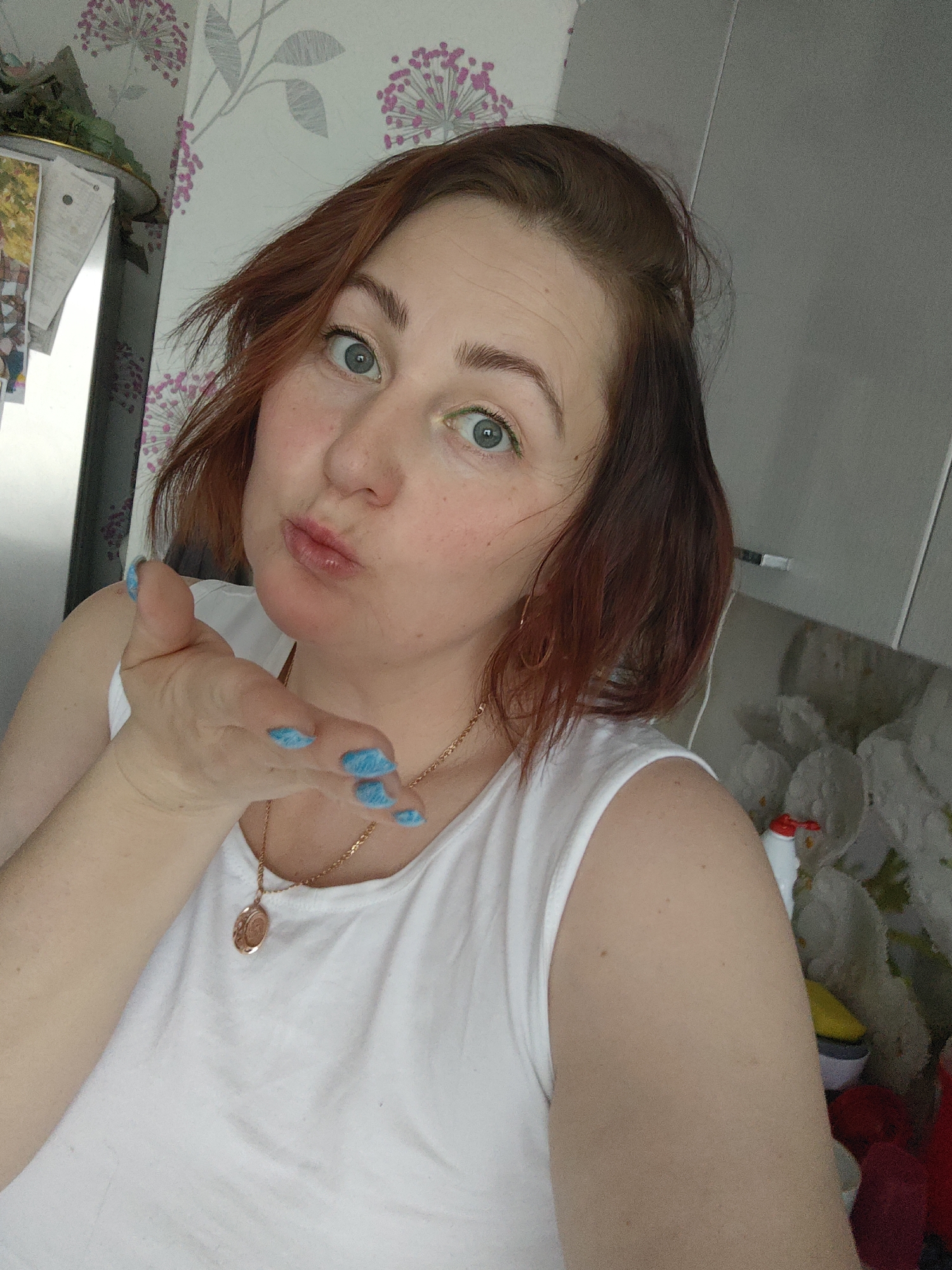 An indoor selfie captures a woman from the chest up, blowing a kiss towards the camera. She has reddish-brown, medium-length hair, and her light blue-green eyes are wide open, with a faint line of green eyeliner visible on her lower lash line. Her lips are pursed for the kiss, and her left hand is extended forward, displaying vibrant blue, possibly glittery, fingernails. She is wearing a simple white sleeveless top and a gold-colored necklace with a circular pendant.
The background reveals a home interior, likely a kitchen or living area. The wall behind her features a light-colored wallpaper with a recurring pattern of pink and gray floral or dandelion-like designs. To the left of the woman, a silver appliance, possibly a refrigerator, is visible with several papers or notes attached to its side. To the right, a plain gray cabinet or wall section is present, along with a glimpse of various colorful household items, including a bottle with a red cap, on a surface below. The lighting suggests it is daytime. The woman's pose conveys a playful and affectionate emotion. No discernible text is visible in the image. The location context for this interior scene is Markovskii, Russia.