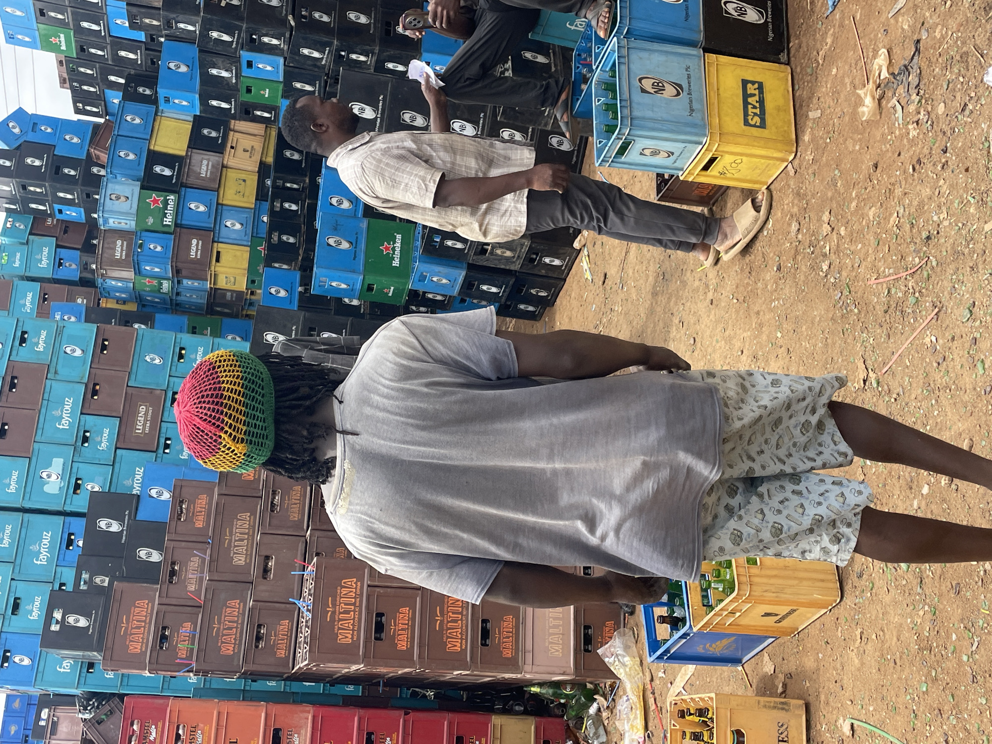 The image shows an outdoor scene in what appears to be a commercial area, possibly in or around Bukuru, Nigeria. A wall of stacked plastic crates dominates the backdrop, labeled with brands like "Maltina," "Fayrouz," "Heineken," "Legend Extra Stout," "Star," and "Nigerian Breweries Plc," indicating a beverage distribution or storage area. Two men are in the foreground. One man, facing away from the camera, wears a rasta-colored hat, a light gray t-shirt, and patterned shorts. The second man, further away, faces the wall of crates and seems to be holding a piece of paper; he is wearing a buttoned shirt and darker pants. In the foreground, there are crates of beverages on the ground, and the ground is covered with dirt and debris.