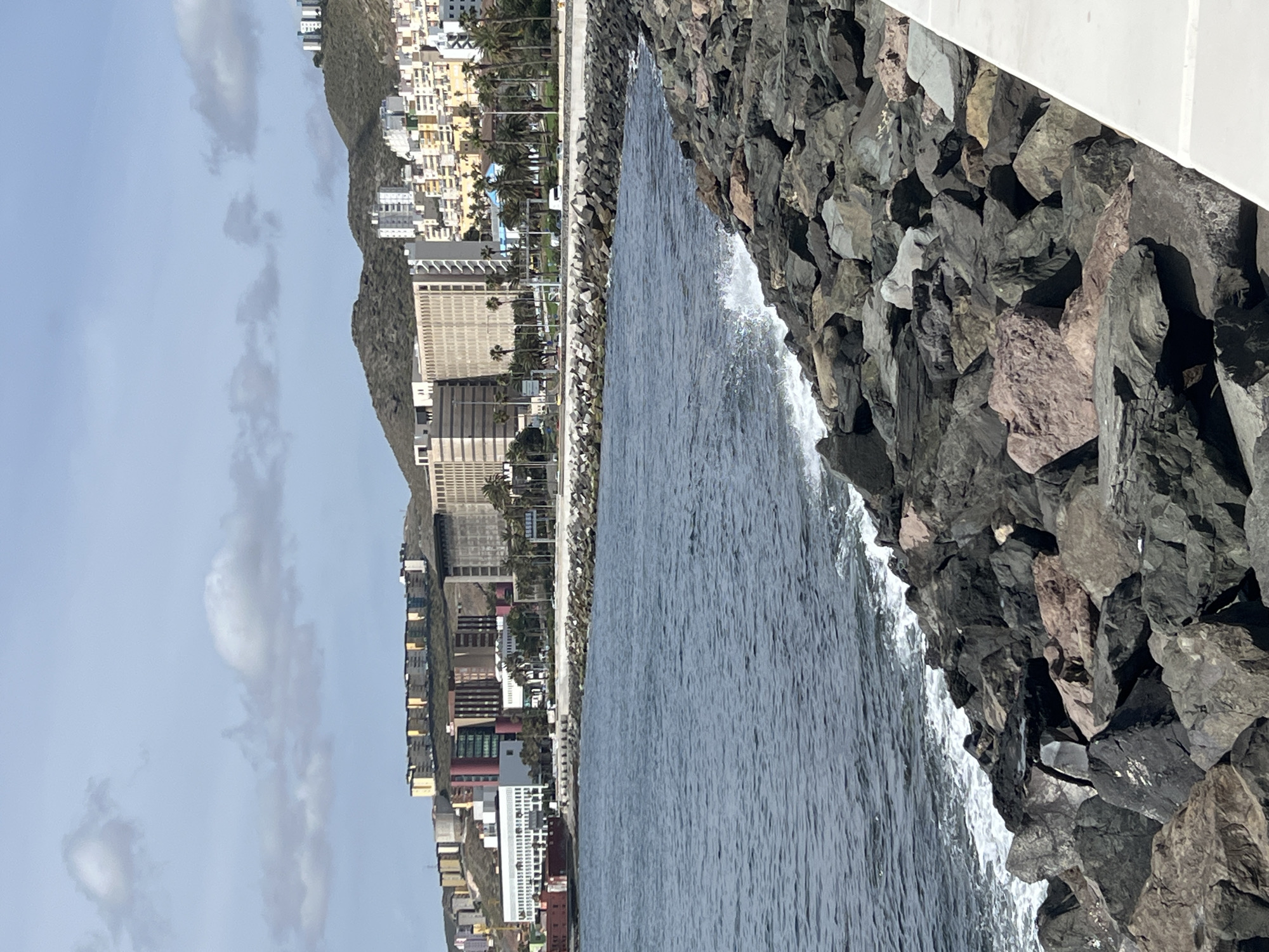 The image shows a waterfront scene in Las Palmas, Spain. In the foreground are large, dark-colored rocks lining the shore, with water surging around them. A white wall or barrier is also present in the foreground. Looking inland, there are densely packed buildings, some with multiple stories and varied colors. Taller buildings, possibly hotels or office towers, are interspersed among the smaller structures. A mountain range rises up behind the city, and the sky above is partly cloudy.