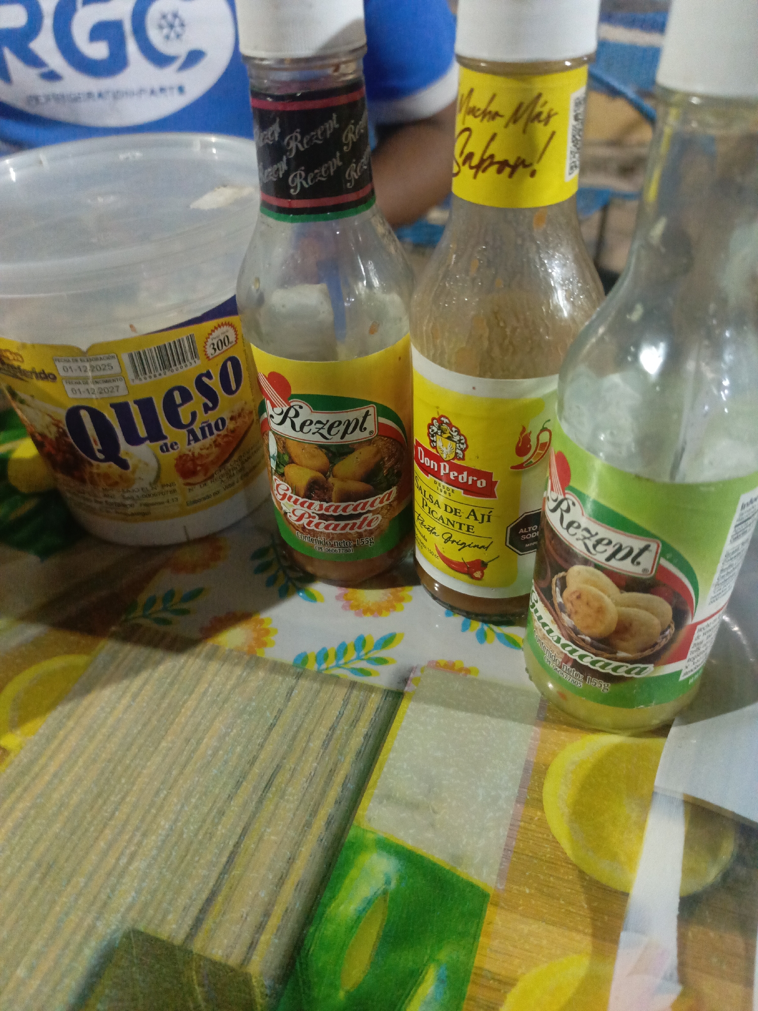 The scene depicts a tabletop setting with several food items. There is a plastic container of "Queso de Año" cheese with a production date of 01-12-2025 and an expiration date of 01-12-2027. Three bottles of sauce are arranged nearby, two labeled "Rezept Guasacaca Picante" and one labeled "Don Pedro Salsa de Aji Picante". A partially visible blue shirt, with the text "RGC" is present, and a second individual can be seen seated in the background. The location is identified as La Tagua, Venezuela. The tabletop has a yellow and green floral and striped design.