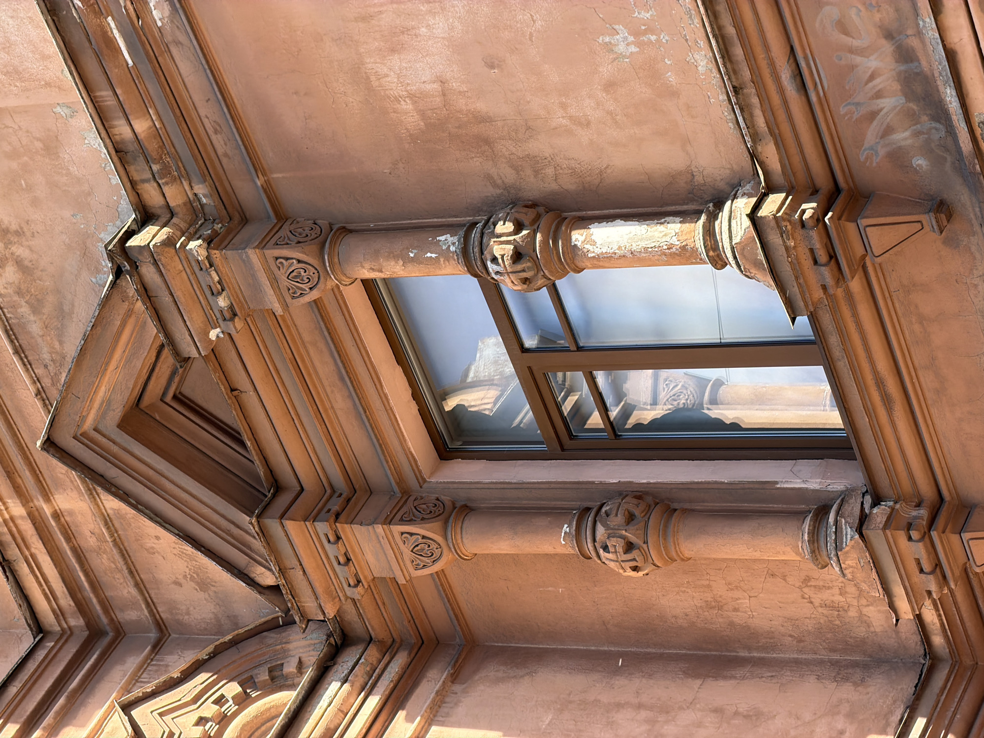 The image shows a close-up view of a segment of a building facade in Saint Petersburg, Russia. The building features ornate architectural details, including carved columns flanking a window and decorative molding along the upper facade. The color of the building is a faded terracotta or salmon pink, with areas of peeling paint and visible cracks in the stucco, suggesting age and wear. The window itself is a modern replacement with brown frames, reflecting the sky and surrounding architecture. The overall impression is one of historical architecture that has experienced some degradation over time.