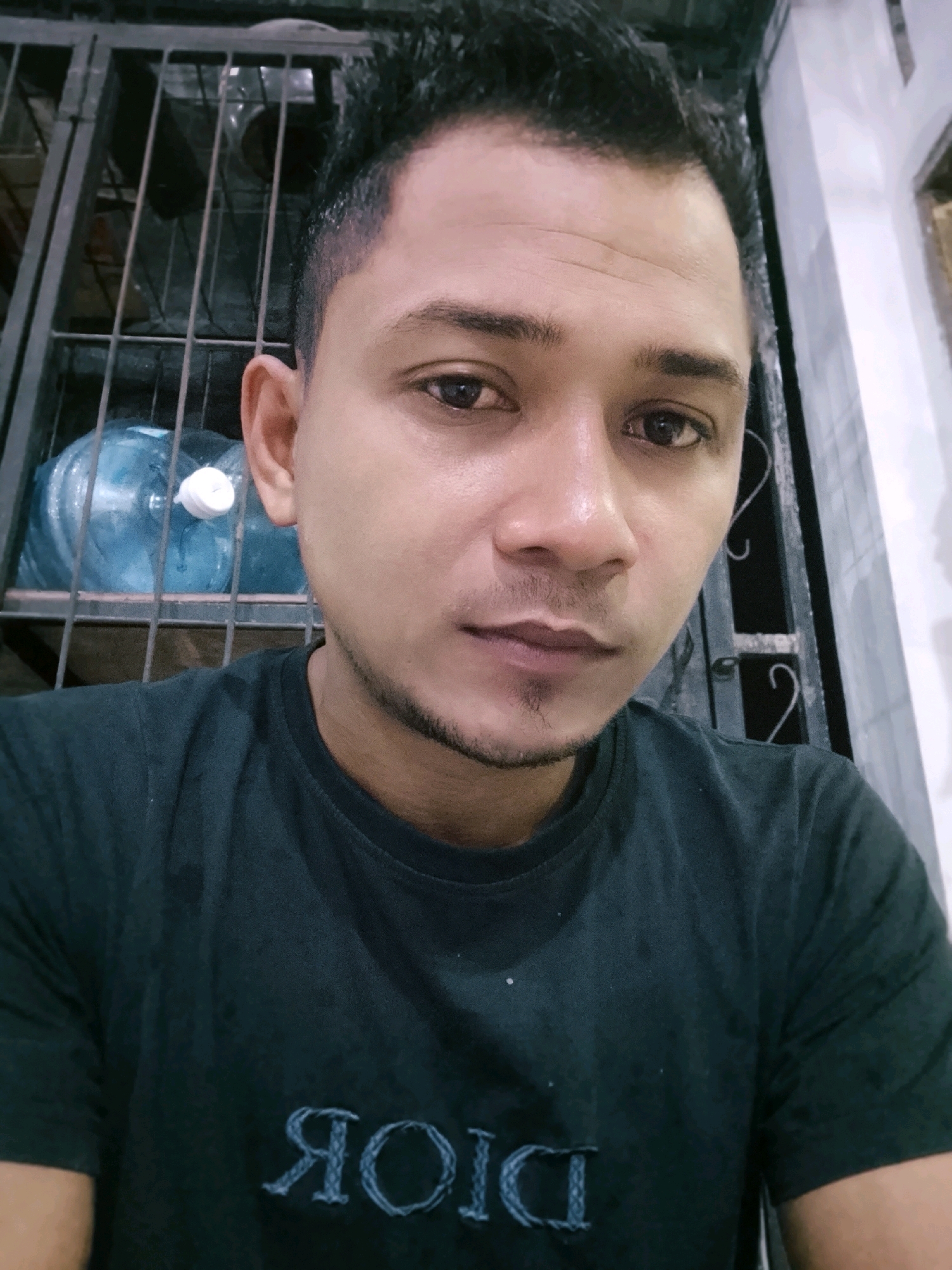 A self-portrait is captured of a man in his early to mid-twenties, positioned in the foreground. He has short, dark, styled hair, dark eyes, and a light brown complexion. His mouth is closed, and he appears to be looking directly at the camera. He is wearing a dark green t-shirt with an embroidered logo on the chest that appears to read "DIOR" in reverse. In the background, behind the man, there is a metal cage or grate. Within the confines of this cage, a large blue plastic water jug with a white cap is visible. To the right of the man, a white wall or structure is partially visible, along with a sliver of a dark window. The overall lighting suggests an indoor setting, possibly at dusk or in an area with limited natural light. The image is taken at a close-up perspective, focusing primarily on the man's face and upper body. The location is specified as Dayahlhoksukon Barat, Indonesia, though there are no explicit visual cues in the image to confirm this specific location beyond the textual information provided.